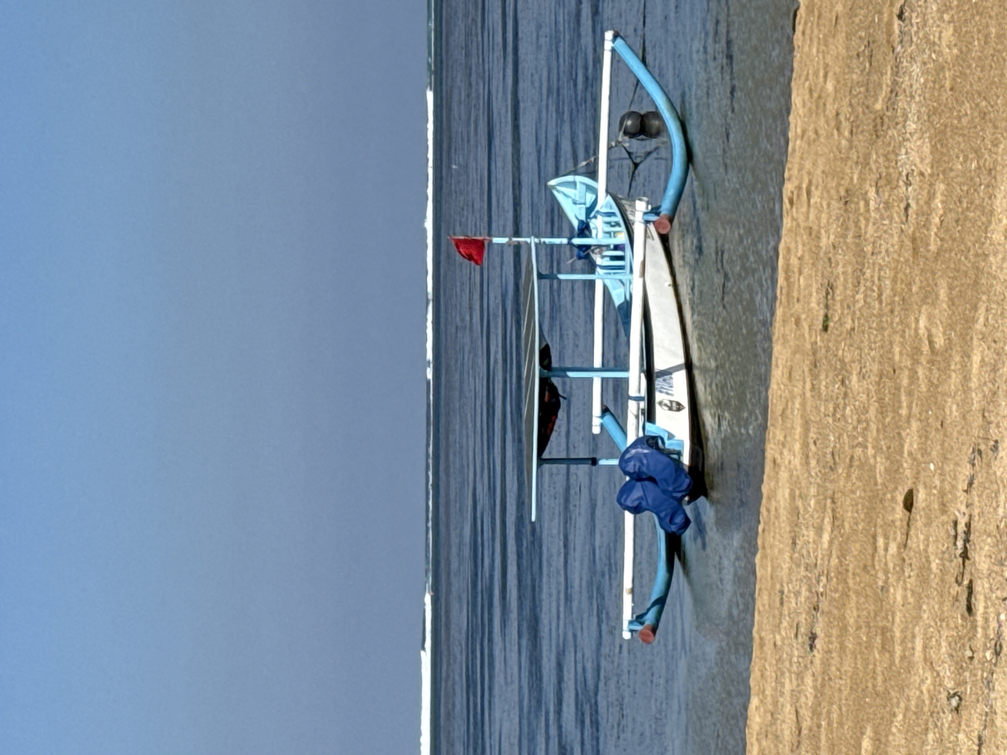 A traditional Indonesian fishing boat with outriggers rests in shallow, calm water near a sandy beach. The boat is painted white with blue accents and features a canopy structure. Two blue cushions are visible on the deck. A small red flag flies from a pole at the stern. In the background, a gentle wave line breaks on the horizon under a clear blue sky. The sand in the foreground is light brown and textured, with some small debris visible. The image appears to be taken in or near Denpasar, Indonesia, likely at a coastal area known for its beaches and fishing activities.