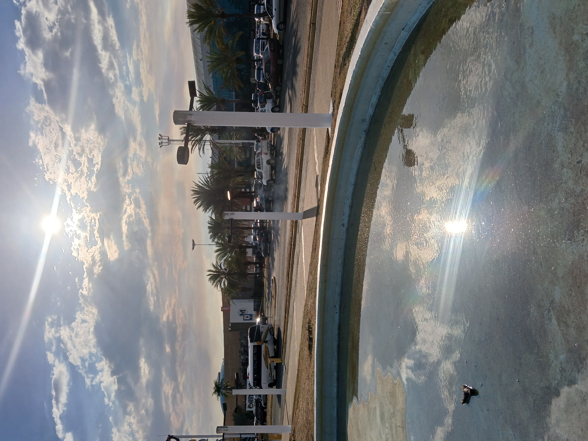 The image captures a sunny day in Ciudad Guayana, Venezuela. A circular fountain occupies the foreground, its water reflecting the sun and sky. Adjacent to the fountain is a road with parked vehicles, and palm trees line the street. In the background is a large building with a reflective glass facade. The sky is filled with clouds.
