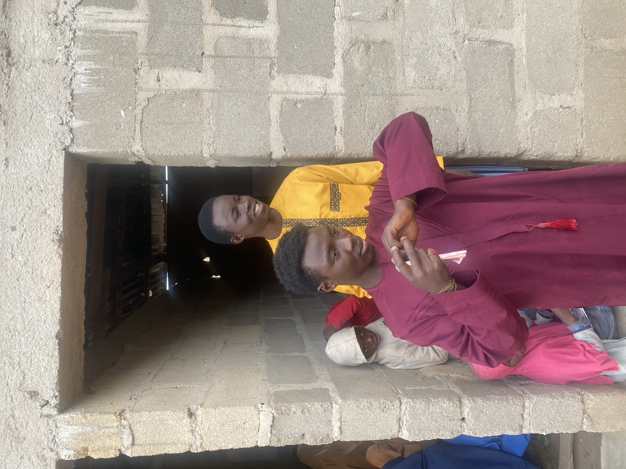 In Jos, Nigeria, two young men stand in a doorway of a building constructed from grey concrete blocks. The man in the foreground, wearing a maroon robe with a tassel, is looking down and appears to be handling something small, possibly money, with his hands. Behind him and slightly to the left, another young man, wearing a yellow robe with decorative trim, is smiling and looking to his left. Partially visible behind the man in maroon, a child in a beige hooded garment peeks out, and to the left of the child, another person in a pink headscarf is visible. The doorway leads into a dark interior where a corrugated metal roof is visible. The overall scene suggests a casual, possibly domestic, setting.