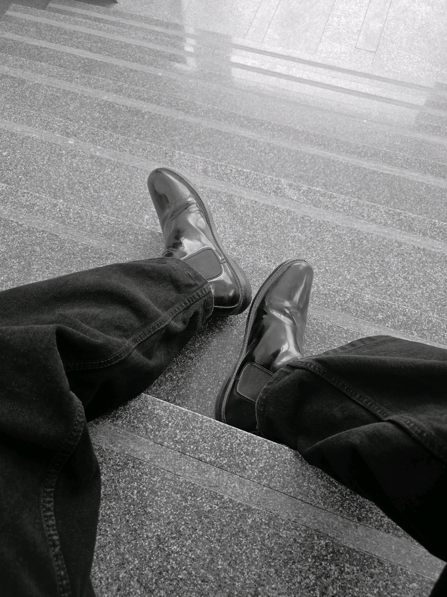A first-person perspective shot shows a pair of shiny black Chelsea boots and dark-colored pants against a speckled gray stone staircase. The boots are well-polished and the pants are creased, suggesting a potentially formal or semi-formal setting. The staircase has several steps visible, with textured stone surfaces and darker, possibly metal, risers. The lighting is somewhat diffused, creating subtle shadows. The scene is devoid of any other people or urban elements, focusing solely on the footwear and lower body of the individual. There is no information in the image to suggest it is located in Ittagalapura, India.