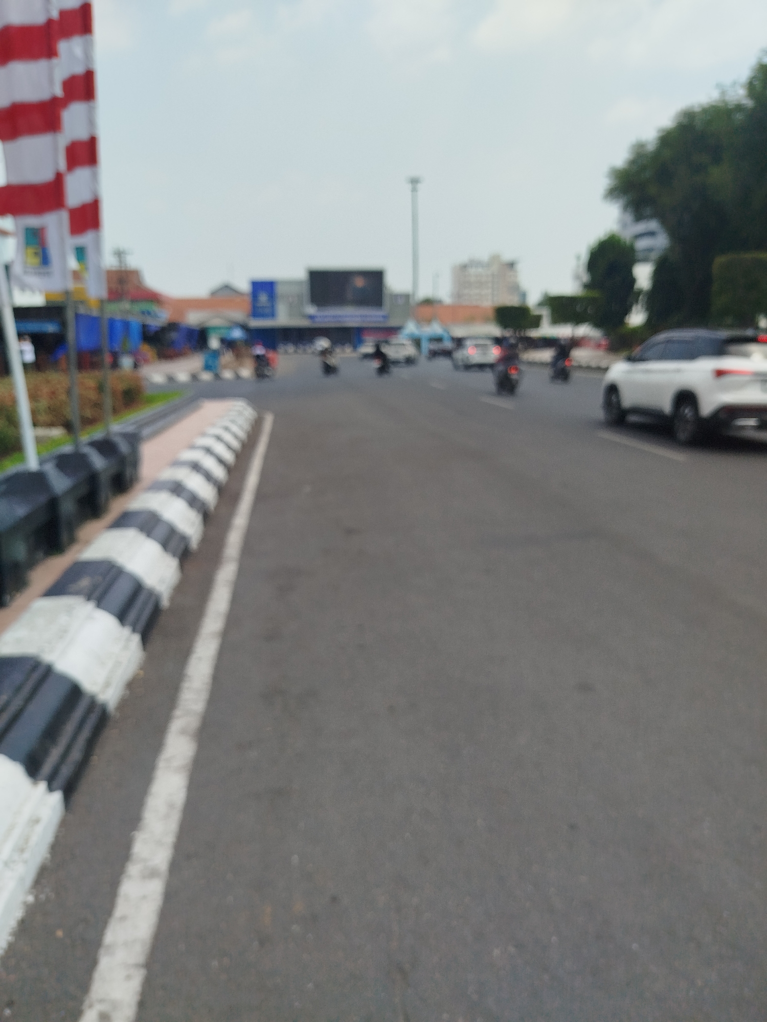 The image captures a blurred scene of a street in Semarang, Indonesia. In the foreground, a black and white striped speed bump lines the edge of the asphalt road, with a white lane marker visible to its right. The background reveals a street with several vehicles, including a white car and multiple motorcycles, in motion. Buildings with blue awnings and signs are visible along the street, alongside a large digital billboard. To the left, a tall pole with red and white striped flags is partially visible. The sky is overcast with light clouds.