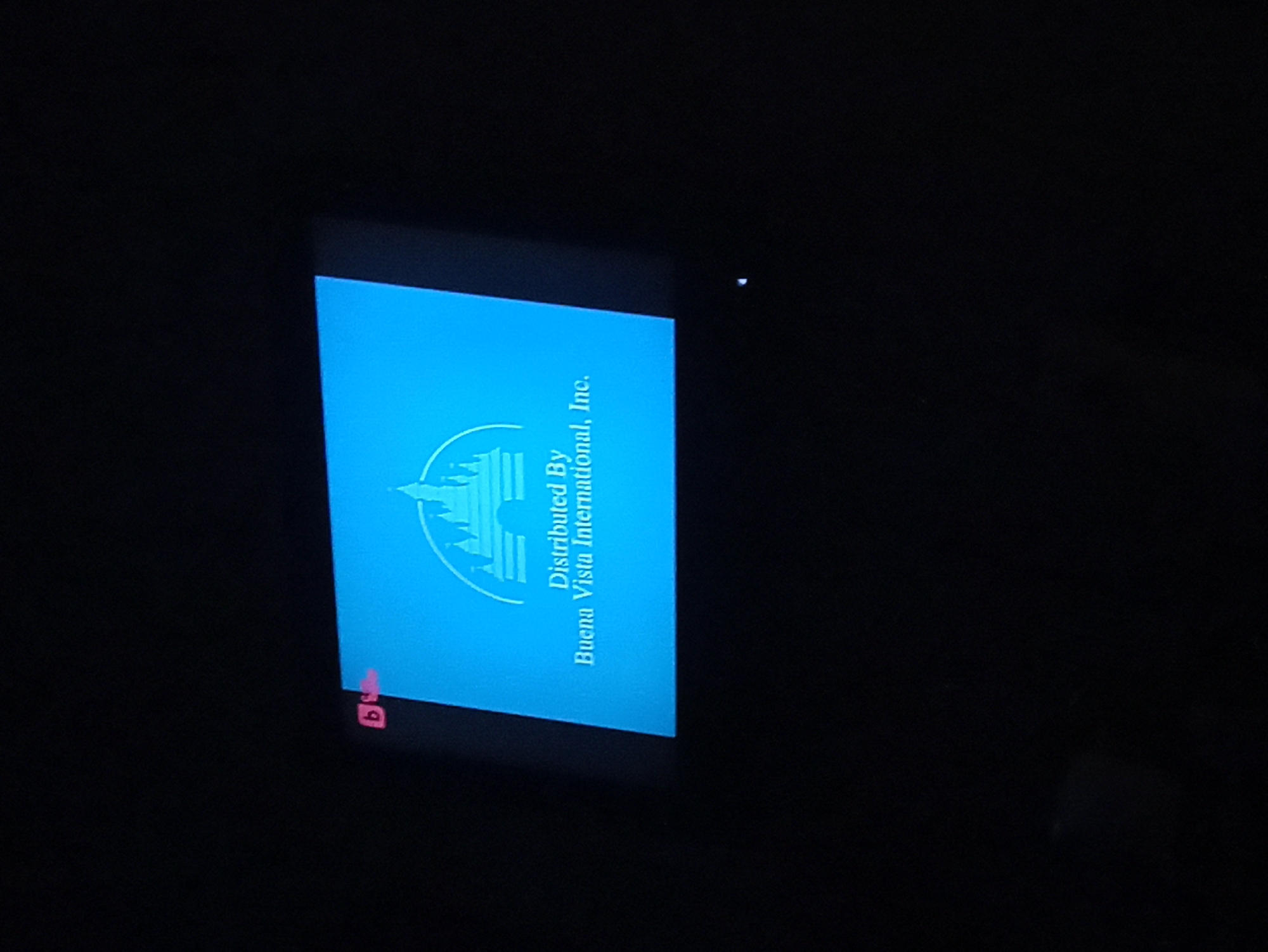 The image shows a blue screen displaying the Buena Vista International, Inc. logo. The surrounding environment is completely dark. A small, red symbol is visible at the bottom left corner of the screen. The location is unspecified.