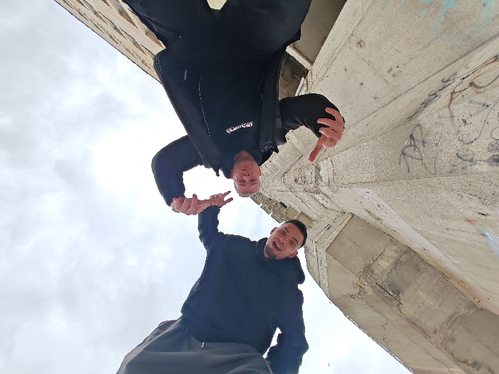 The image displays two males against a large, textured concrete structure under an overcast sky. The photograph is taken from a low-angle perspective, causing one subject to appear inverted.
The subject in the lower portion of the frame, positioned on the right, is a male with short dark hair and a beard. He wears a black hooded sweatshirt and dark grey sweatpants. He is looking directly at the camera with his mouth open. His right hand is raised, with two fingers extended in a gesture.
The second subject, positioned in the upper portion of the frame and inverted, is a male with short light-colored hair. He wears a black jacket with a white logo visible on the left chest area and dark pants. His right hand (viewer's left) makes a downward pointing gesture, while his left hand (viewer's right) is open with fingers splayed. His facial expression is neutral.
The background features a massive, angular concrete wall or pillar, exhibiting a rough, weathered texture consistent with brutalist architecture. Patches of graffiti, including blue and black markings, are present on the concrete, particularly on the right side of the frame. An expanse of bright, cloudy sky occupies the upper-left and central portions of the background.
This concrete structure is identified as a component of the Monument to 1300 Years of Bulgaria, located in Shumen, Bulgaria.