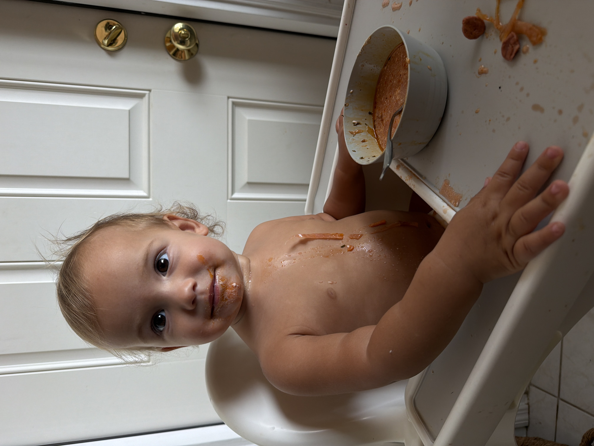 A toddler sits in a high chair with a bowl of food and a spoon. The child has brown eyes, light hair, and food smeared on their face and chest. The high chair is white, and the tray is covered with food. A white door with gold hardware is visible in the background. The location is not discernible.