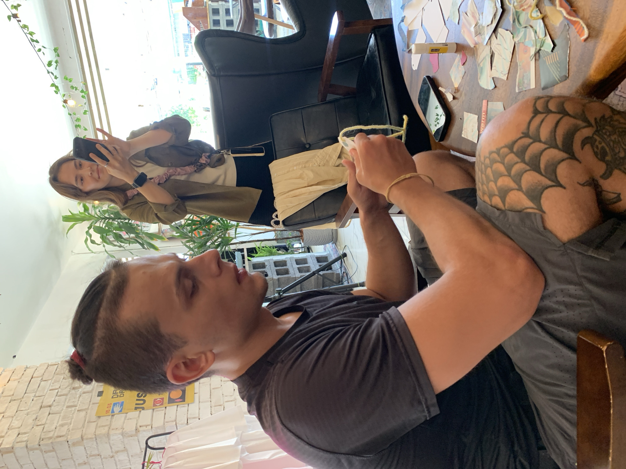 In Baan Bo Phut, Thailand, a man with a man bun is seated at a wooden table, focusing on a yellow piece of yarn he is manipulating with his hands. He is wearing a dark t-shirt and grey shorts, and has a spiderweb tattoo on his thigh. To his right, a woman stands holding a smartphone, appearing to take a photo or video. She is wearing a green jacket over a white t-shirt and has a necklace with a pink beaded strap. Behind them, a large black leather armchair is visible, with a folded beige cloth draped over its back. The environment suggests an indoor setting, possibly a cafe or workshop, with plants and decorative lights visible in the background. The table is cluttered with small pieces of paper, a glue stick, and a smartphone.