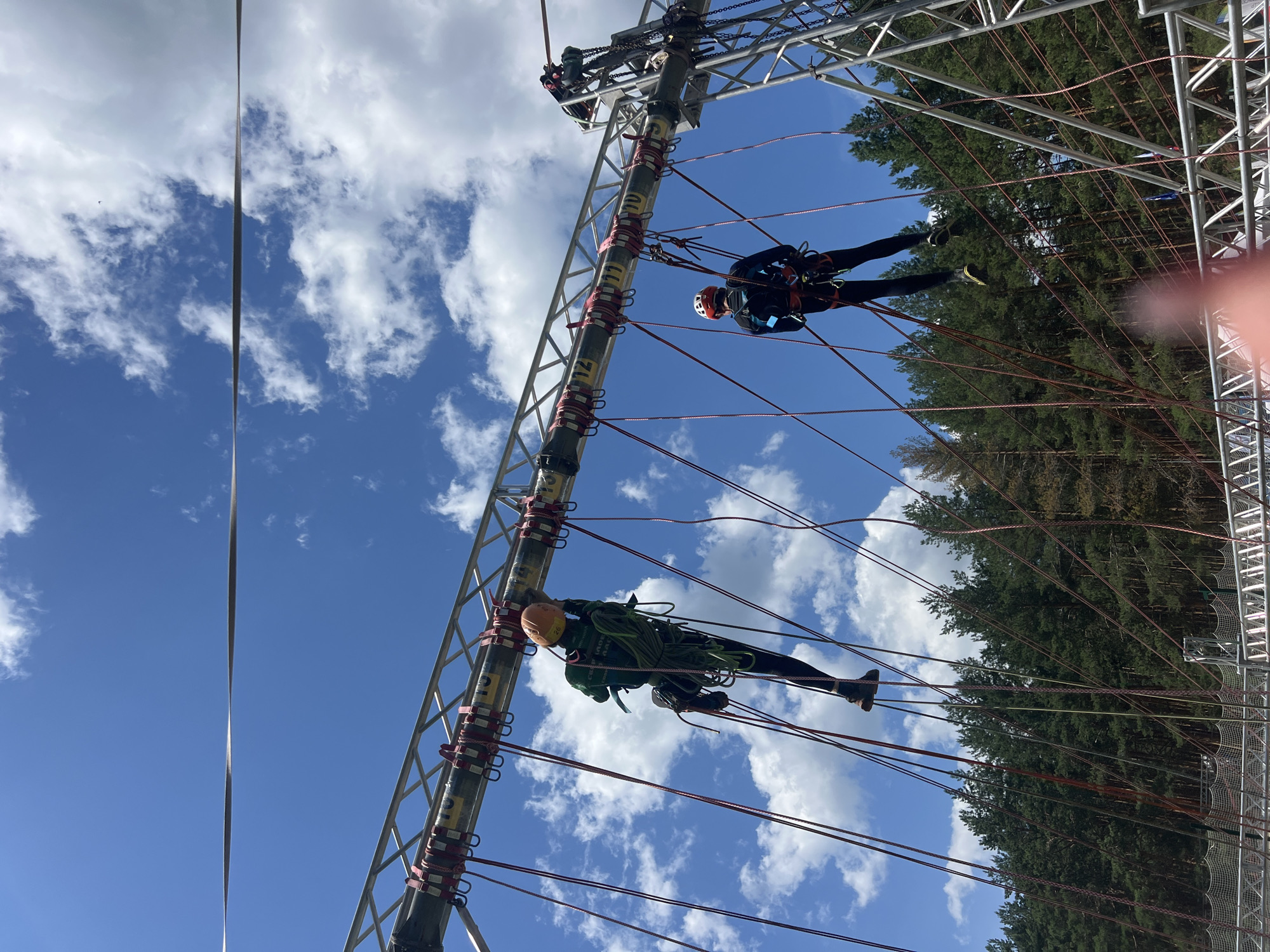 Two individuals, wearing helmets and harnesses, are navigating an aerial obstacle course. The course consists of a metal superstructure with numerous ropes. One person, dressed in green, is positioned on the left, facing away from the camera, suspended by ropes. The other individual, in a dark blue suit, is on the right, also suspended by ropes and facing away from the camera. Both are positioned against a backdrop of a bright blue sky with scattered white clouds and a dense forest of evergreen trees. The scene appears to be an outdoor adventure park, likely in Penza, Russia, given the context provided.
