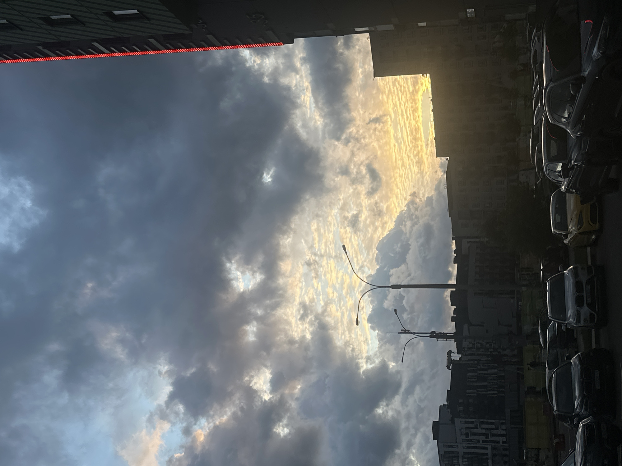 The image depicts a view in Бараўлянскі сельскі Савет, Belarus, showing a sky filled with dark, grey storm clouds interspersed with areas of bright, golden sunlight. The sky is framed by buildings on either side, with a building on the top left featuring a strip of red lights along its edge. On the right side, parked cars line the street. Streetlights are visible in the middle of the frame, silhouetted against the bright sky. In the distance, buildings of varying heights are visible.