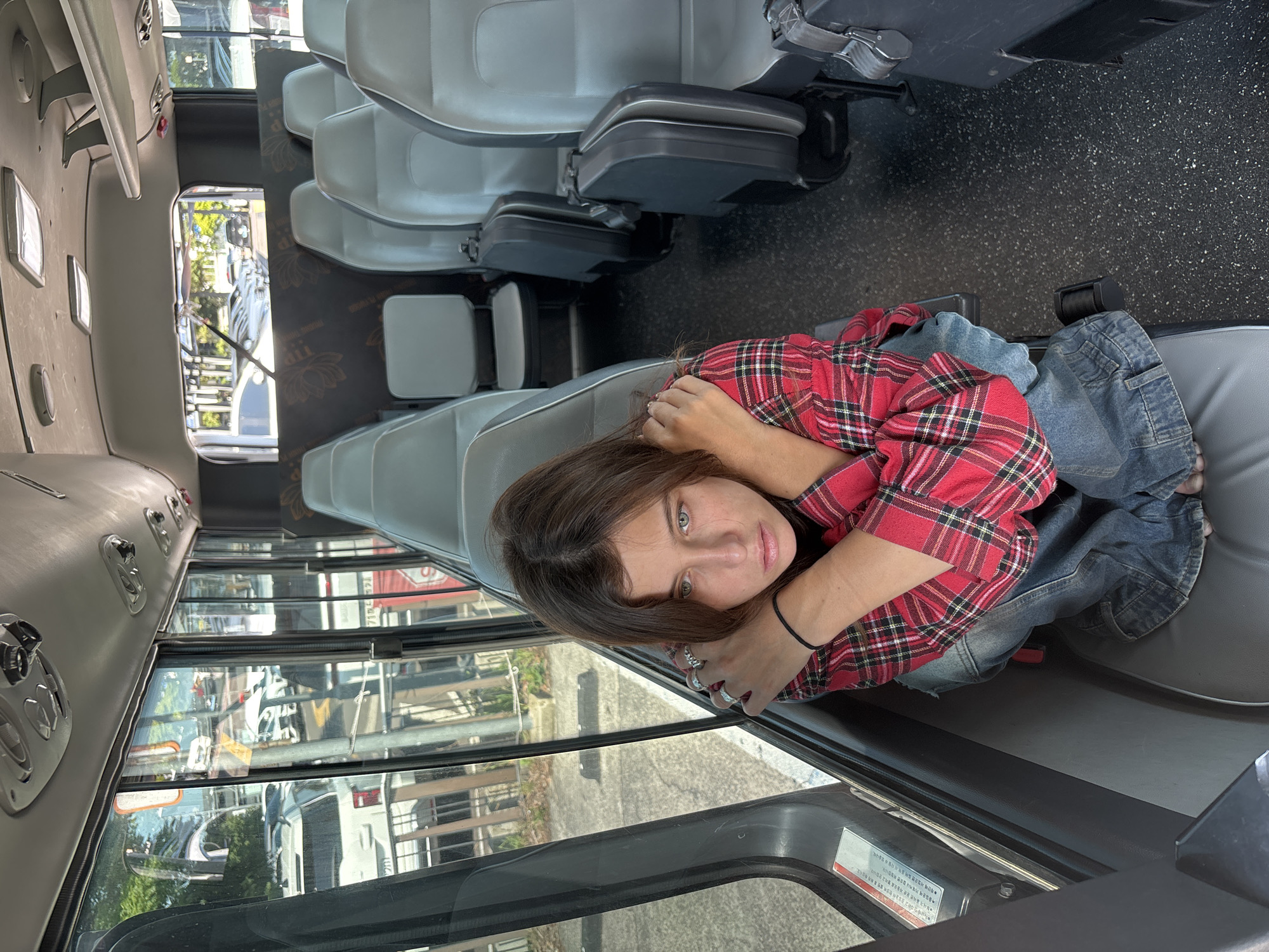 A young woman with green eyes and brown hair is lying down on a bus seat, looking at the camera. She is wearing a red and black plaid shirt and blue jeans. Her arms are around her head, and she has several rings on her fingers. The interior of the bus is visible, with rows of gray seats and windows that show a blurred outdoor scene. The setting appears to be a sunny day, with natural light coming through the windows. The background outside the windows suggests an urban or suburban environment with trees and buildings. A few pieces of text are visible on a sticker on the interior of the bus, but they are not legible. The overall mood is relaxed and candid.