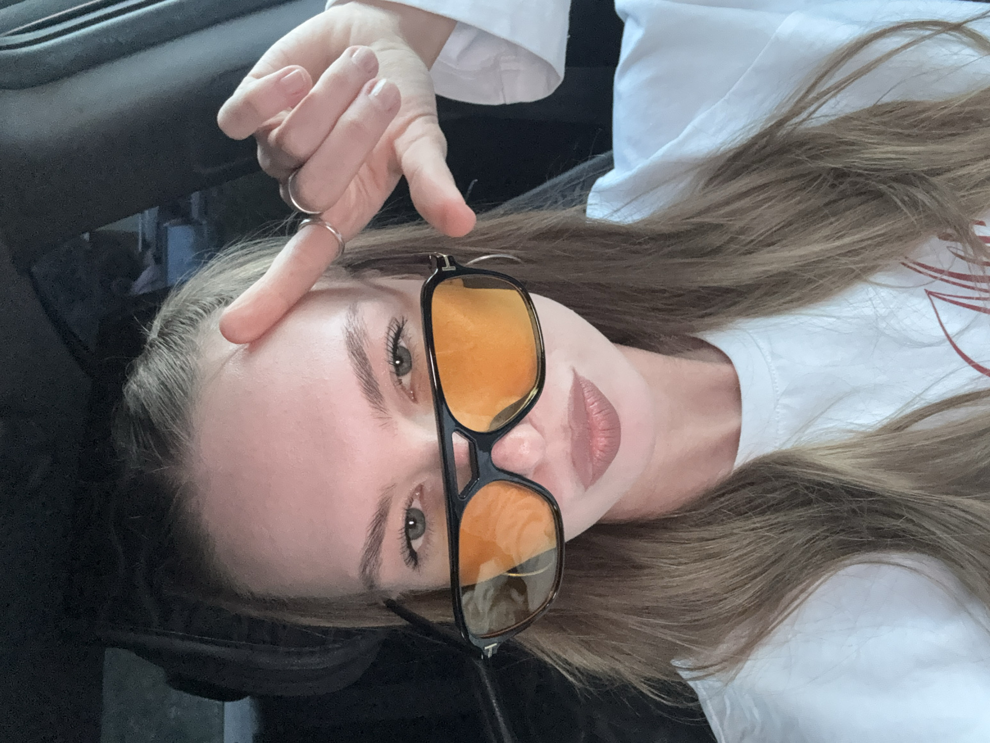 The image shows a close-up of a young woman wearing large, dark-rimmed sunglasses with orange lenses. She is looking at the camera with her lips slightly pursed. Her right hand is raised, with her index finger touching her forehead and her other fingers curled. She is wearing a white t-shirt and has long, light brown hair. The background is dark and appears to be the interior of a car, suggesting the photo was taken there. There are no specific landmarks or environmental clues that would indicate the city of Perm, Russia.