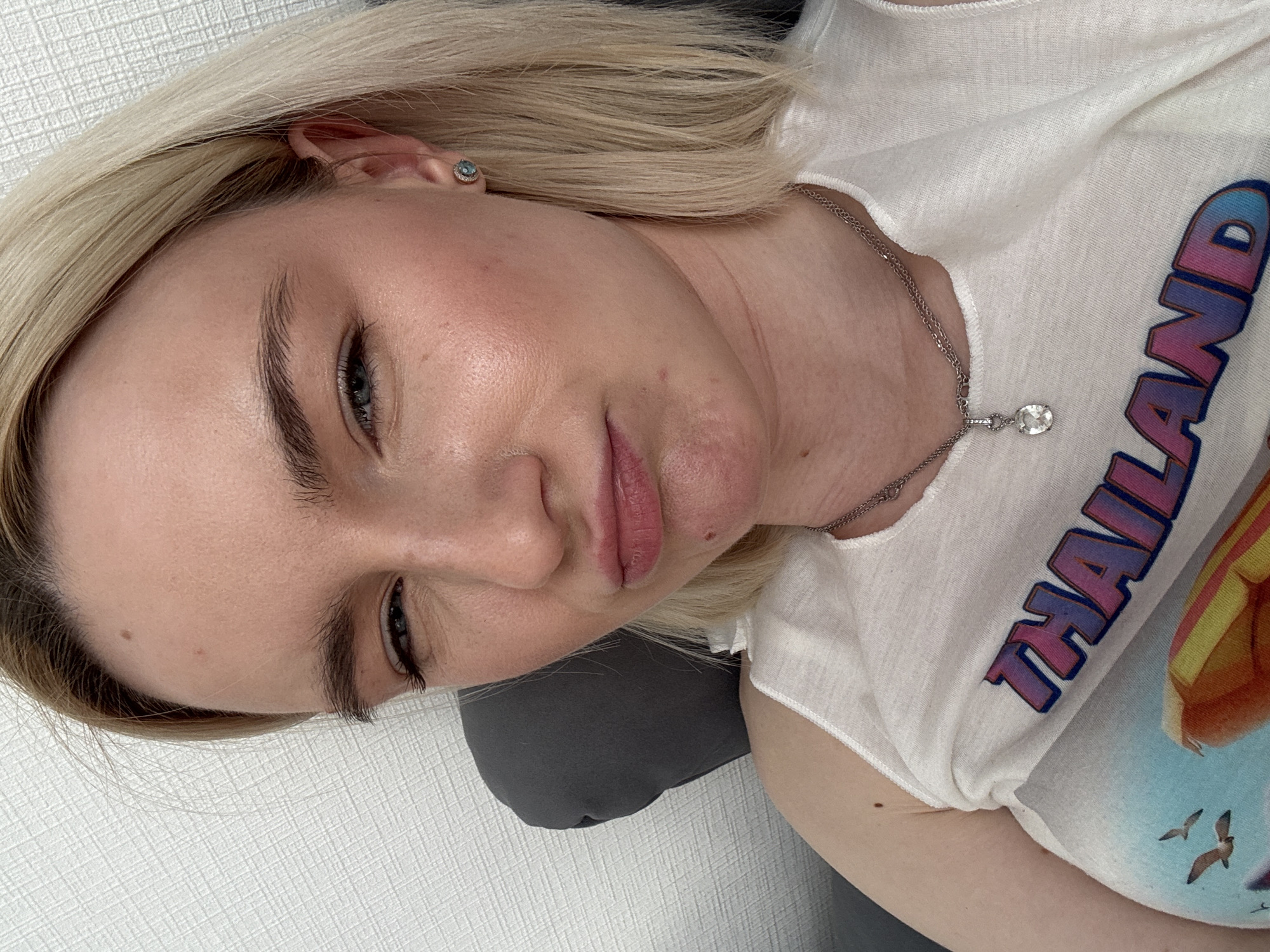 A light-skinned woman with blonde hair is depicted from the chest up, looking downward. She is wearing a white t-shirt with the word "THAILAND" printed on it, and a silver necklace with a pendant. She also has a blue gemstone earring on her left ear. The background features a white textured wall and a grey cushion or pillow. There is no evidence in the image that the location is Vsevolozhsk, Russia.