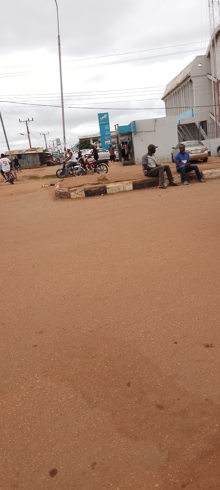 The image depicts a street scene in Gboko, Nigeria, on a cloudy day. In the foreground, a dirt road with reddish-brown soil dominates the lower half of the frame. A raised curb, painted with alternating black and white stripes, separates the road from the area where people are gathered.
Several people are present. On the curb, two men are seated. The man on the left wears a grey shirt and dark pants, while the man on the right is in a blue t-shirt with white lettering and dark pants. Further down the curb, a group of individuals appears to be standing near a building.
On the street, there are motorcycles and people riding them. One motorcycle, a blue scooter, is being ridden by a man in a white tank top. Another motorcycle, red, is parked behind it, with a person on it. To the left, a person on a white motorcycle with the number "51" on the back is moving away from the camera. Several other motorcycles are visible in the background, suggesting active transportation in the area.
Behind the people and vehicles, a light-colored, modern building with vertical architectural elements is visible. A blue and white sign with an emblem resembling a horse and the text "UnionBank" and "ATM" is also prominent, indicating a bank branch. To the left of the bank, a simpler, corrugated metal structure is present. Overhead, power lines and a tall lamppost are visible against the overcast sky. The overall impression is that of a busy urban intersection.