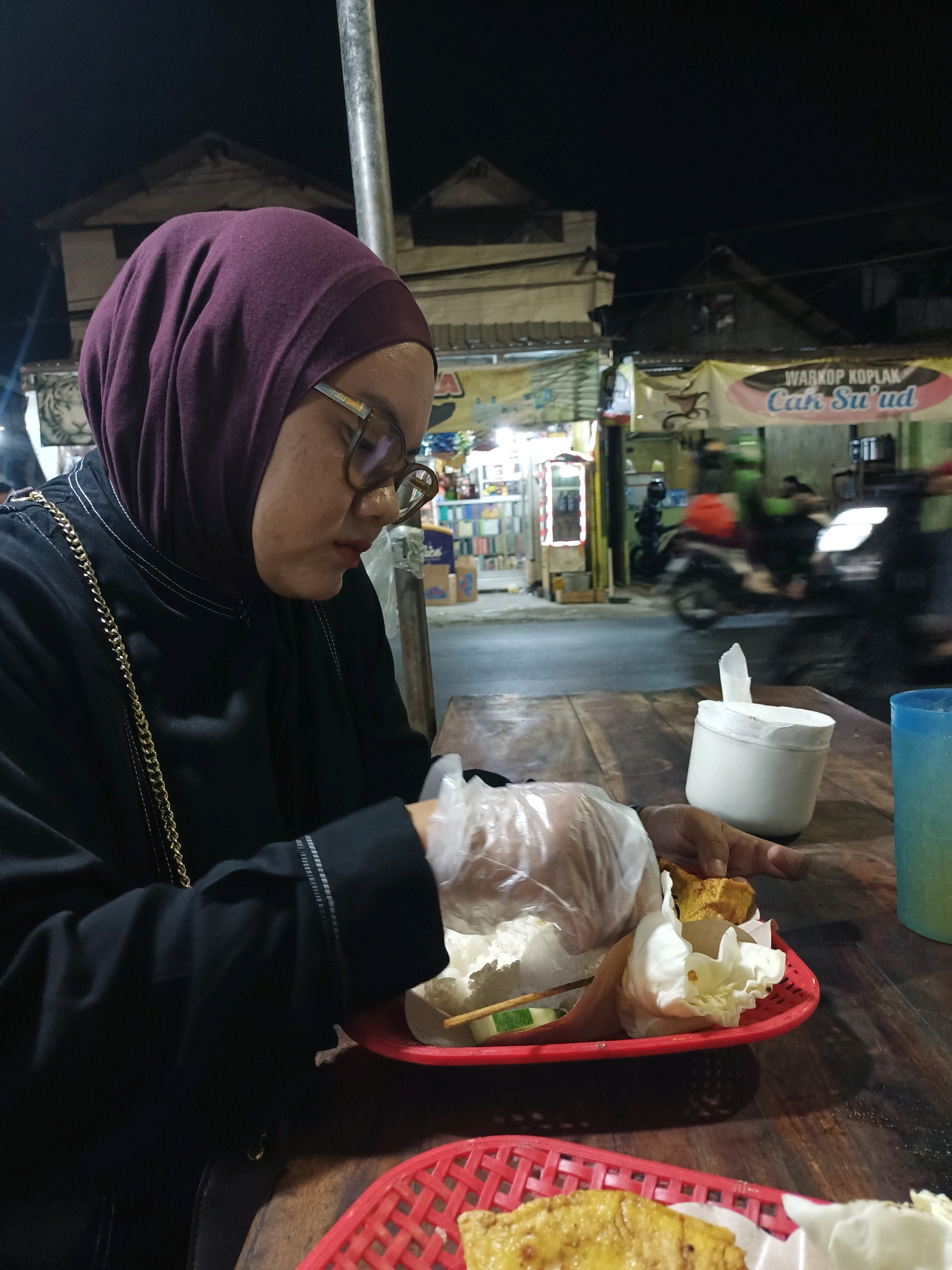 A woman wearing a dark hijab and glasses is seated at a wooden table outdoors at night, preparing to eat from a red plastic tray containing food wrapped in plastic. She is wearing gloves and appears to be holding a piece of food. The background shows a street scene with buildings and parked motorcycles, suggesting a roadside eatery or warung. A banner with text reading "WARKOP KOPIAH Cak Su'ud" is visible. The scene is dimly lit, with artificial lights illuminating the area. The overall atmosphere suggests a casual dining experience.