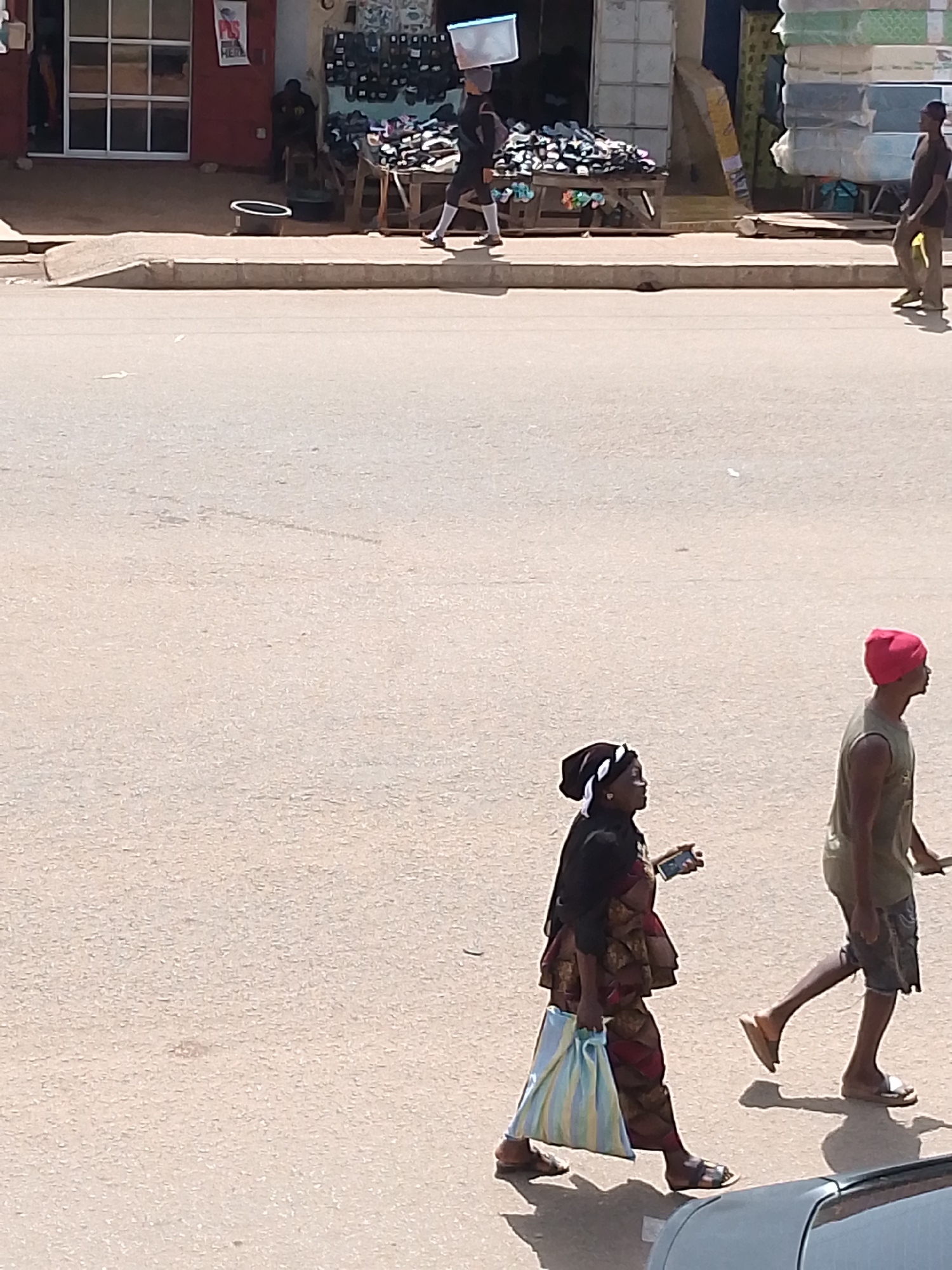 In the city of Bukuru, Nigeria, an outdoor scene unfolds on a sunlit street. Two individuals are captured mid-stride on the pavement, their figures silhouetted against the muted tones of the urban landscape.
To the left, a person walks with a light blue and yellow striped bag held casually in their left hand, their gaze directed forward. They wear a dark garment with a colorful, patterned skirt and sandals. A black and white headscarf covers their hair, and a white band adorns their forehead.
To the right, another person walks briskly, their right arm swinging slightly. They are dressed in a sleeveless olive-green shirt and dark shorts. A vibrant red beanie caps their head, and they wear sandals. The two individuals are walking in the same direction, though at a slight distance from one another.
The background offers glimpses of a bustling market area. Structures with red and white facades line the street, with open doorways revealing displays of goods. A rack overflowing with footwear is visible to the left of the central path. In the distance, stacks of mattresses are piled high, suggesting a commercial district. The street itself is paved with asphalt, showing signs of wear and tear, with subtle markings and scattered debris. The sunlight casts distinct shadows on the ground, indicating a bright, clear day. A parked vehicle, partially visible in the foreground, adds to the everyday nature of the scene.