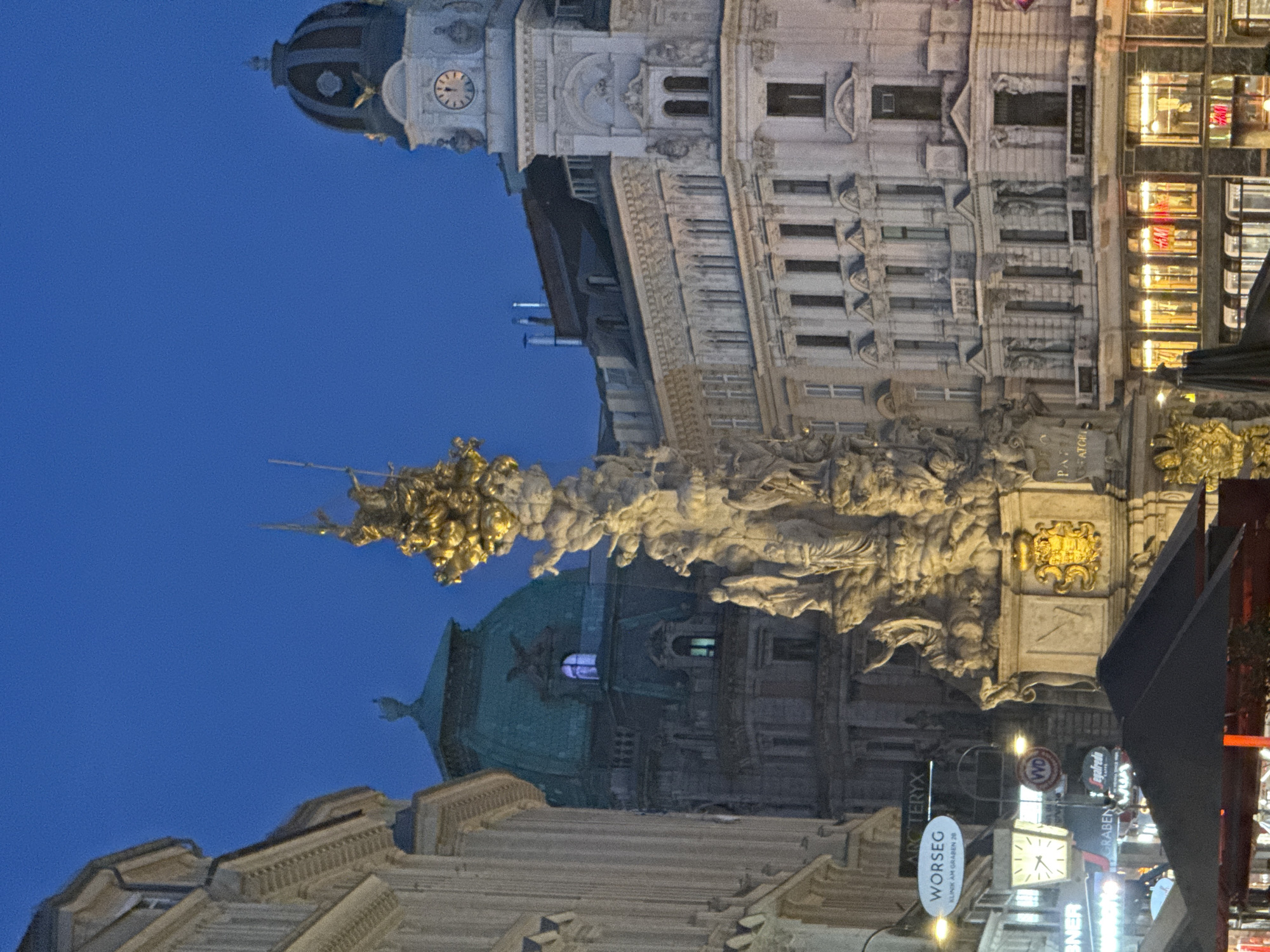 This is a nighttime, rotated, wide-angle shot of a bustling city square in Vienna, Austria. The scene is dominated by ornate historical architecture, including a tall building with a clock tower and a domed structure. A prominent element is a large, detailed sculpture or monument featuring numerous figures, gilded elements, and what appears to be a statue of an angel or winged figure at its apex.
The activity seems to be the evening ambiance of a public space. Streetlights and illuminated shop windows cast a warm glow, indicating it's after dark. There are several signs visible with text, including "WORSEG KLINIK AM GRABEN 26", "TERRYX", and "WD". Other signs suggest commercial establishments, with one appearing to be for H&M. The sky is a deep, clear blue, suggesting fair weather. The overall mood is one of historical grandeur meeting modern urban life.