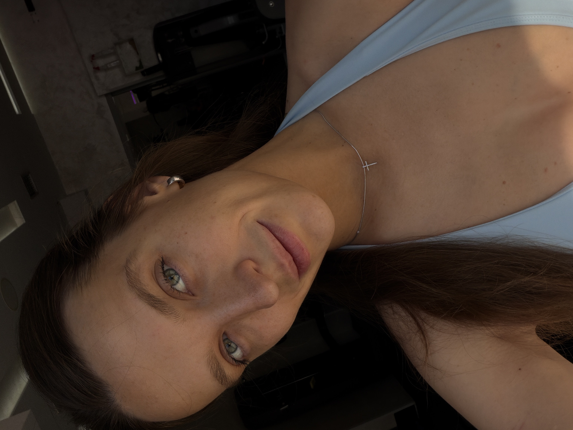 This is a close-up selfie of a young woman indoors. She has long brown hair, light skin with freckles, and green eyes. She is wearing a light blue halter-neck garment and a delicate silver necklace with a small cross pendant. Her left ear is adorned with a silver hoop earring. The background is dark and out of focus, suggesting an indoor environment, possibly a gym or a private space. There is no discernible indication of her location in Budva, Montenegro.