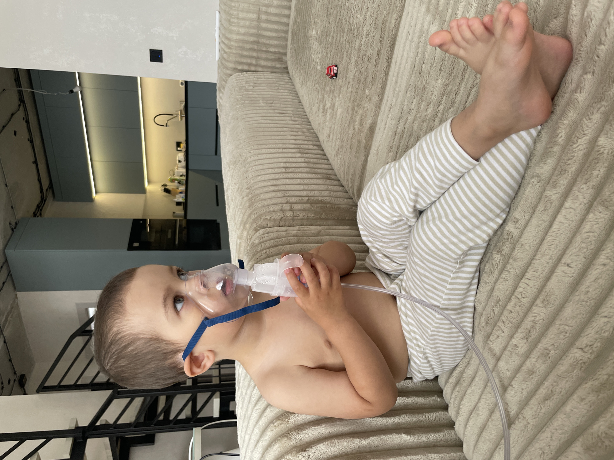 A young boy is sitting on a plush, beige couch in an indoor setting, undergoing a nebulizer treatment. He is shirtless and wearing horizontally striped pants. The nebulizer mask covers his nose and mouth, and he holds the device in his hands. A clear tube connects the nebulizer to an unseen machine. The background reveals a modern kitchen with light blue cabinets and accent lighting, along with a staircase. A small red toy car is visible on the couch beside him. The scene takes place inside an apartment.