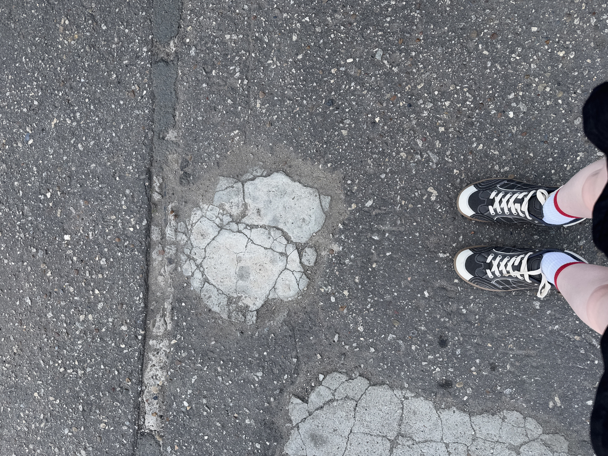 A high-angle shot depicts a section of cracked asphalt pavement. A repair patch, characterized by a lighter-colored and heavily fractured concrete-like material, is visible near the center. To the right of the frame, a pair of feet wearing black and white sneakers with white laces and red-striped white socks is present. The person wearing the shoes is also wearing a black garment. The location, Balezino, Russia, cannot be verified from this image.