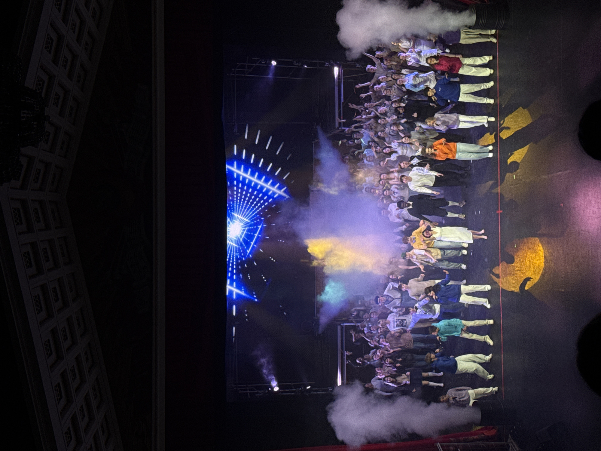 Here is a description of the image.
The image shows a large group of people on a stage in Perm, Russia. The crowd is facing forward, with some individuals positioned horizontally across the stage, possibly performers or participants in a presentation. Lighting effects are in use, including blue and yellow projections, along with smoke or fog effects. Architectural details of the building's interior are visible along the left edge of the frame.