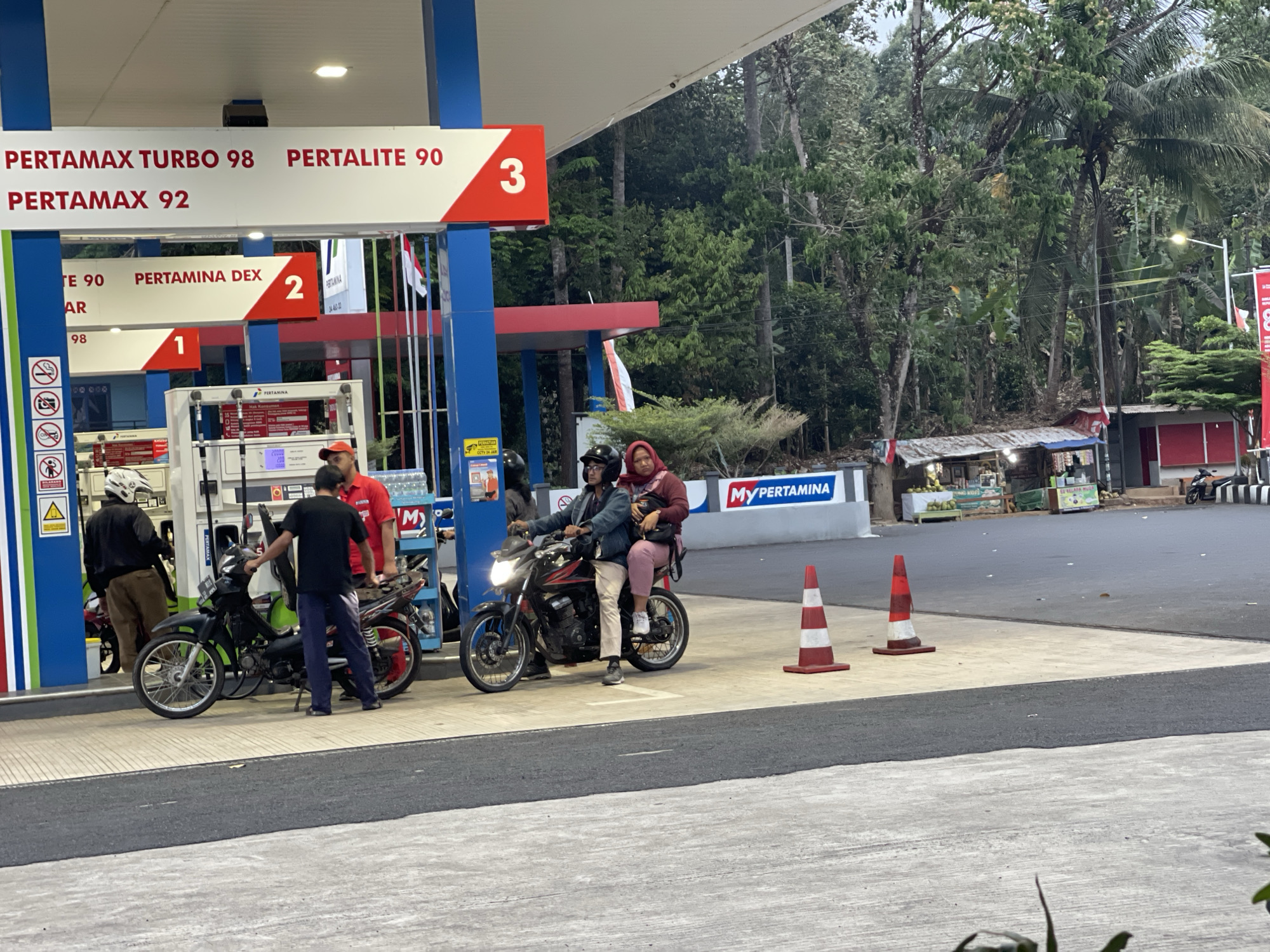 The image depicts a Pertamina gas station in Ciamis, Indonesia, at dusk. In the foreground, several motorcycles and people are visible. Two individuals are seated on a black motorcycle, with the passenger wearing a pink headscarf and the driver wearing a denim jacket and glasses. Another person, wearing a black t-shirt and blue pants, stands next to a black motorcycle, holding onto its handlebars. To the left, another person in a black jacket and white helmet stands near a motorcycle.
The gas station has blue and white dispensers with prominent Pertamina branding. Signs display fuel prices like "PERTAMAX TURBO 98," "PERTALITE 90," and "PERMAX 92." Traffic cones are placed on the asphalt.
In the background, lush green trees and foliage dominate the scene, suggesting a natural or semi-rural environment surrounding the gas station. A small roadside stall or shop with a corrugated metal roof is visible, with a few motorcycles parked nearby. The lighting suggests it's either early morning or late evening.