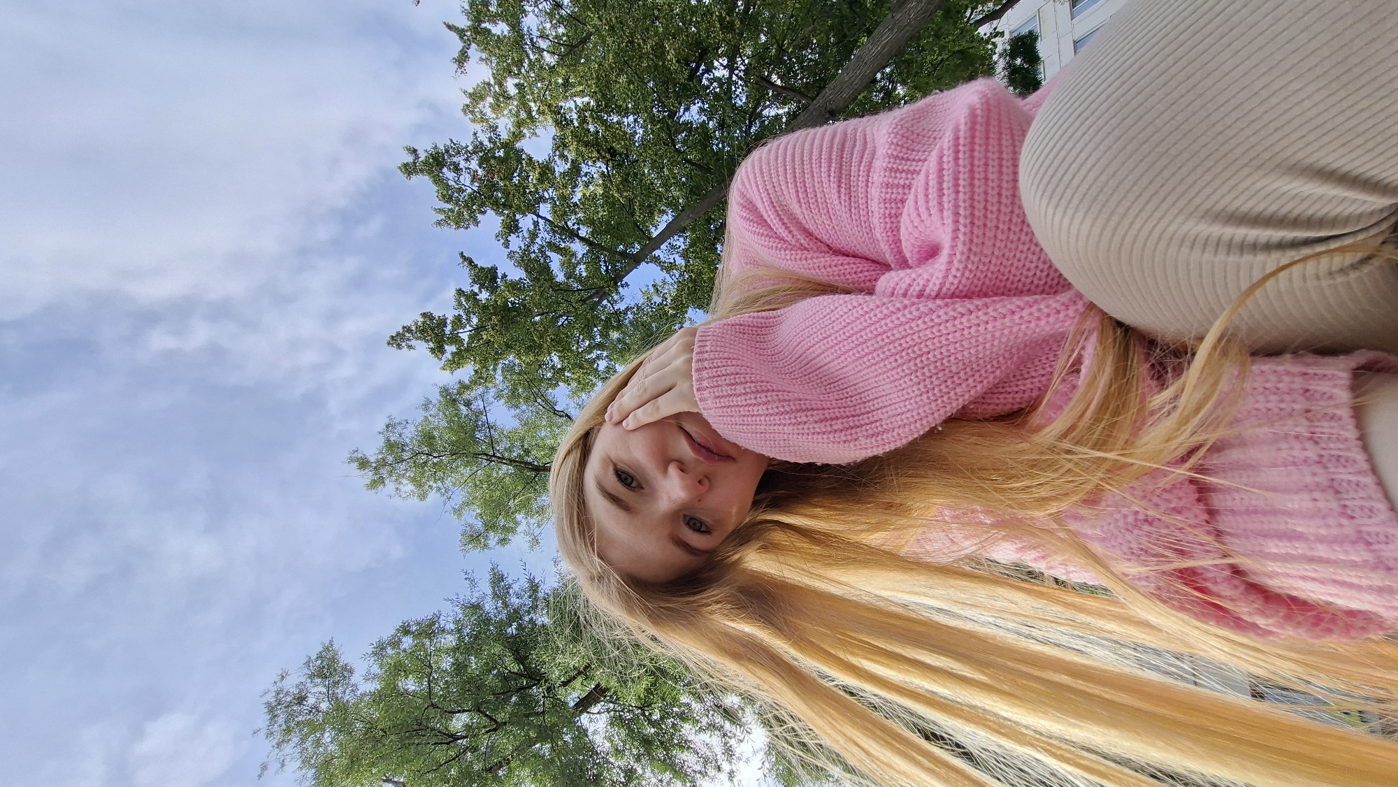 The image is a low-angle, wide shot selfie of a young woman taken outdoors. She has long, blonde hair that cascades down and is partially covered by her shoulder and arm. She is wearing a thick, pink, knitted sweater and light-colored pants. Her right hand is touching her face, partially covering her eye, and she is looking directly at the camera with a slight smile. The background consists of a blue sky with some white, wispy clouds and the green foliage of trees. A portion of a building is visible in the upper right corner. The lighting suggests it is daytime, likely late morning or afternoon, with clear weather. The woman's pose and expression convey a relaxed and cheerful mood. There is no visible text in the image. The scene appears to be set in a park or a natural setting near a building. Given the location context of Chaikovsky, Russia, the image likely depicts a pleasant day in that city.