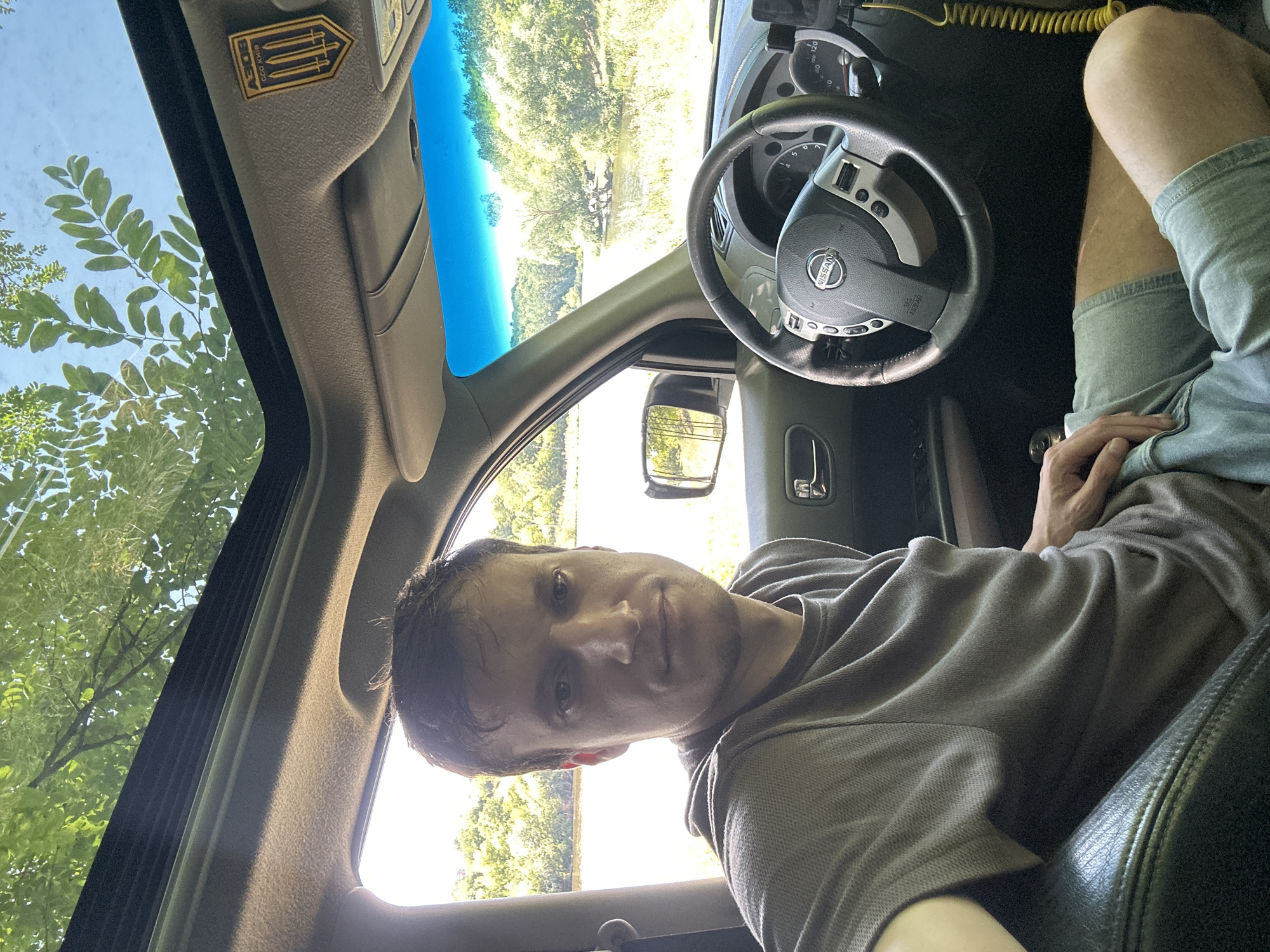 A man sits in the driver's seat of a Nissan X-Trail SUV, with a panoramic sunroof open above him. He is wearing a grey t-shirt and light blue shorts. The vehicle is parked on a dirt road next to a body of water, likely a river or lake, with lush green trees and vegetation lining the banks. The sun is shining brightly, casting shadows. On the visor above the windshield, a patch with three vertical swords within a shield emblem is visible. The backdrop suggests a rural or natural setting, possibly on the outskirts of Makiv, Ukraine, given the presence of the emblem. The man is looking directly at the camera, appearing relaxed. The steering wheel and dashboard of the car are also visible.