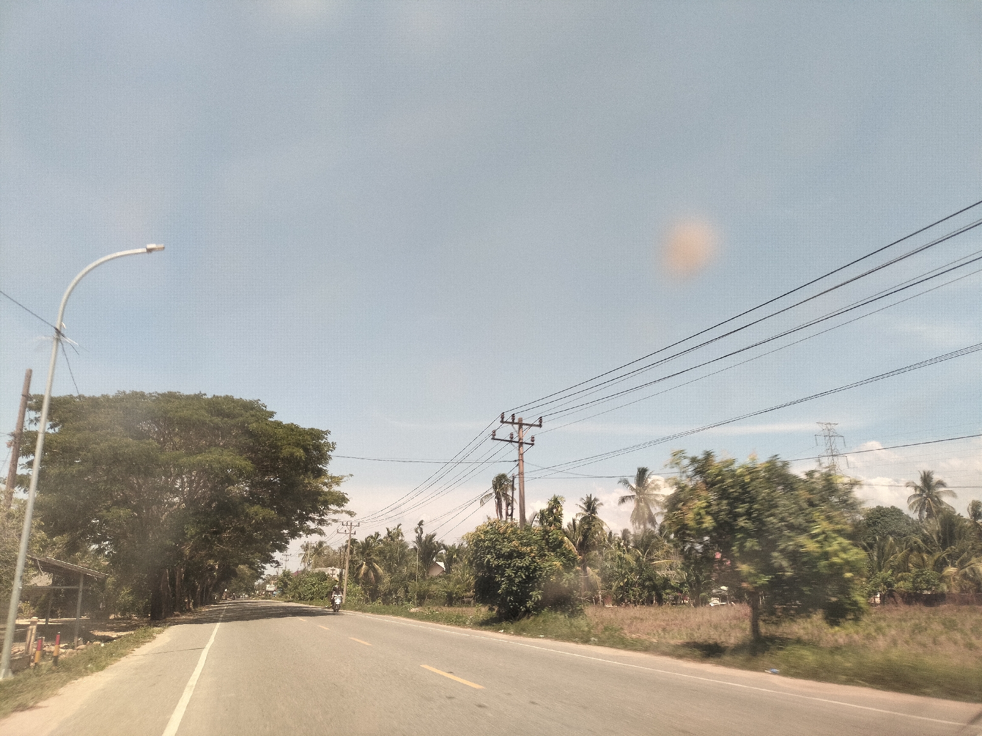 The image depicts a rural road scene in Matangkumbang, Indonesia. The road is asphalt with a yellow center line. A lone motorcycle is traveling in the distance on the left side of the road. Power lines run overhead. On either side of the road are trees, palm trees, vegetation, and occasional buildings, suggesting a tropical or semi-tropical environment. A street light stands on the left side of the road. The sky is clear and light blue.