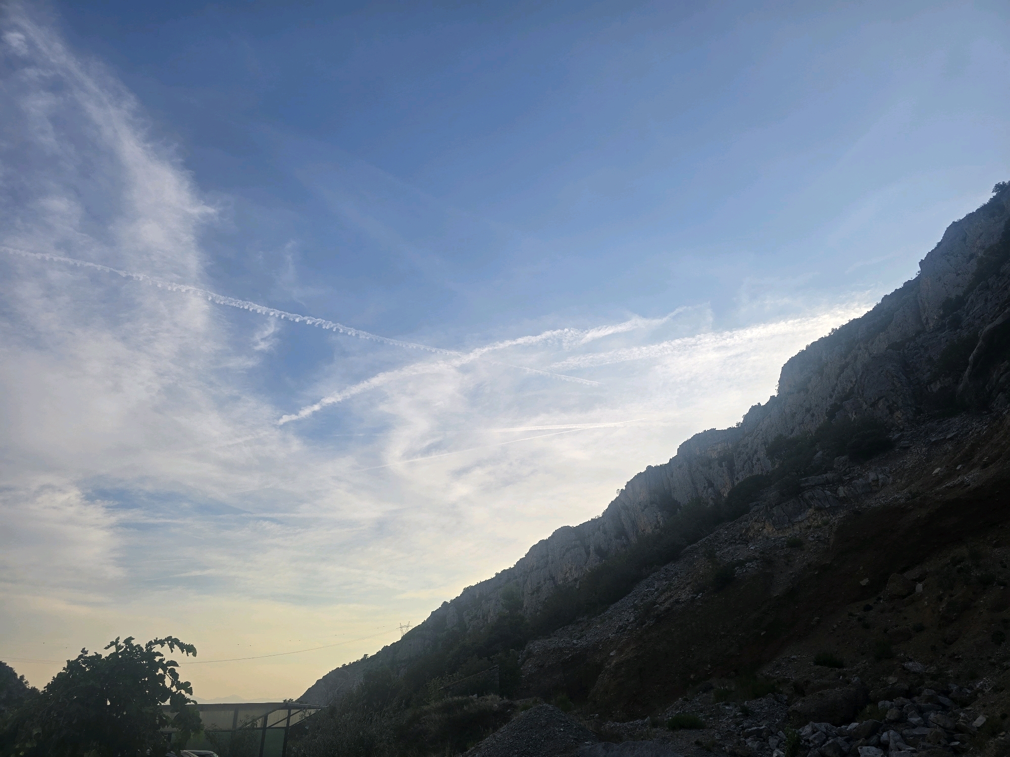 The image depicts a steep, rocky mountainside under a bright blue sky with wispy white clouds and contrails from aircraft. The sun appears to be setting or rising, casting a warm glow on the horizon and creating a gradient of light in the sky. In the foreground, there is some low vegetation and debris, suggesting a natural or possibly industrial area. The overall impression is one of a rugged natural landscape in an outdoor setting. The location is implied to be near Podgorica, Montenegro, though no specific landmarks of the city are visible in this particular frame.