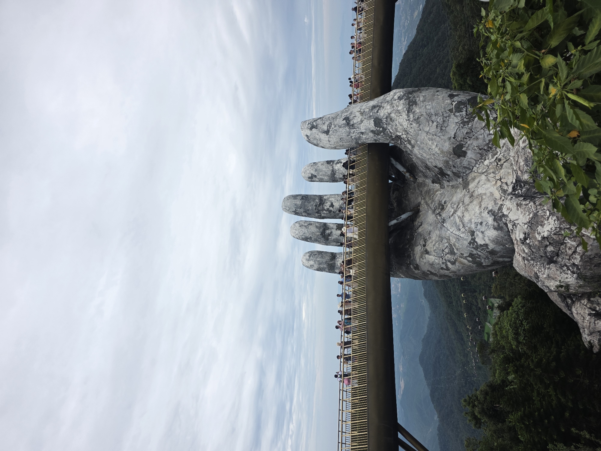 This is a photograph of the Golden Bridge in Đà Nẵng, Vietnam. The bridge is supported by two giant stone hands, and a pathway runs across the bridge. People are walking on the bridge, enjoying the view. The bridge is set against a backdrop of lush green mountains and a cloudy sky. The scene is vibrant and awe-inspiring, showcasing the unique architecture and natural beauty of the location.
