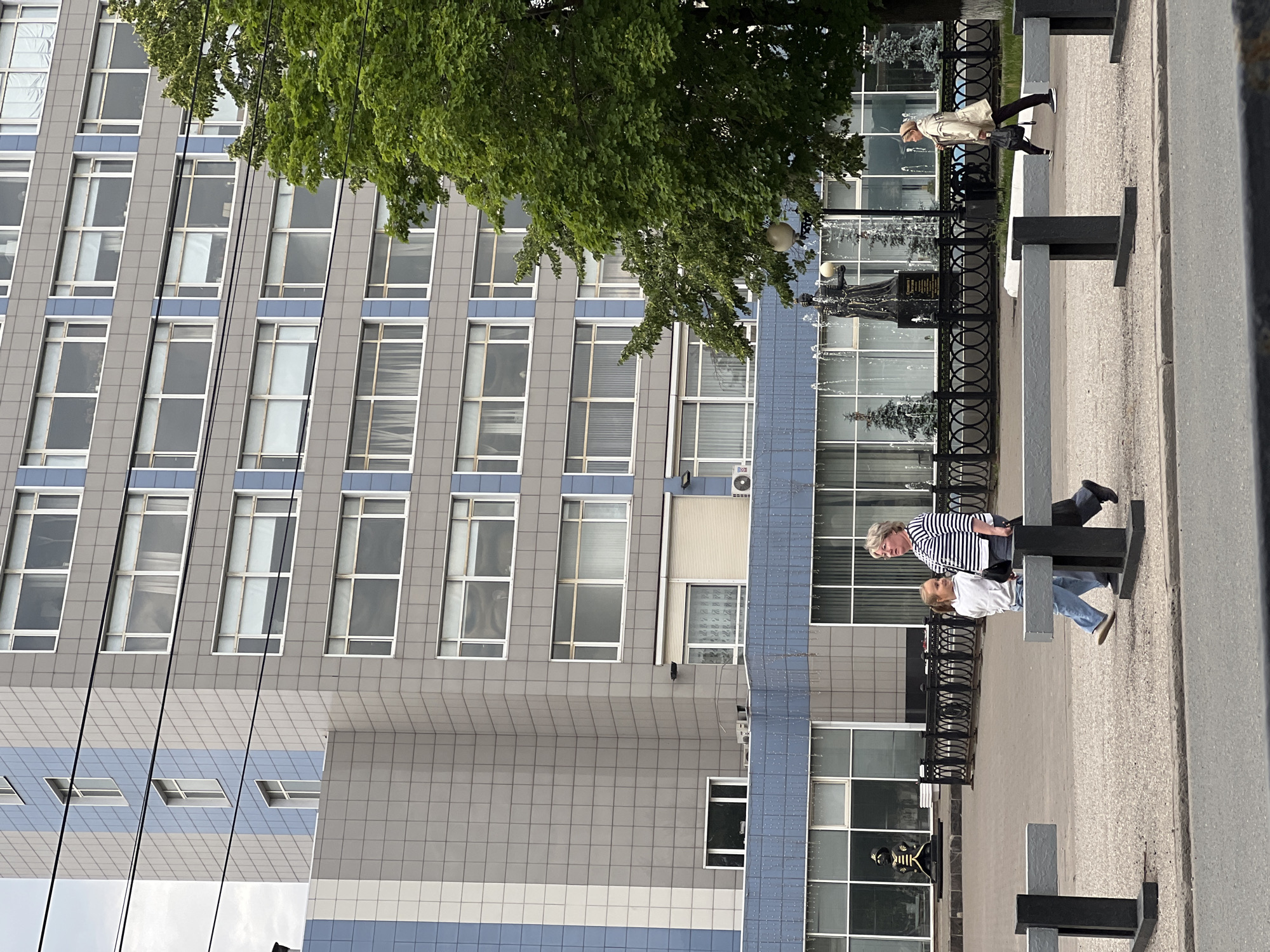 This image captures a street scene in the city of Izhevsk, Russia, with a modern multi-story building dominating the background. The building's facade is a combination of gray and light blue tiled sections, with numerous windows arranged in horizontal rows. A large, mature green tree partially obscures the right side of the building.
In the foreground, two women are walking on a paved walkway. The woman on the left is a younger girl wearing a white hooded sweatshirt and blue jeans. She is walking slightly ahead of the woman on the right, an older woman with blonde hair, wearing a horizontally striped long-sleeved shirt and dark pants. They appear to be walking in the same direction.
Behind them, a decorative black metal fence separates the walkway from a paved area with a fountain. A dark statue, possibly of a historical figure, stands near the fountain, which features multiple water jets. Further to the right, another woman is walking away from the viewer, carrying a dark bag. In the background, beyond the fountain and fence, a large building with extensive glass windows is visible. Sculptural figures, possibly part of the building's design, are also present within the glassed areas.
The sky is overcast, suggesting a cloudy day. Electrical wires are visible running horizontally across the upper portion of the building. The ground in the foreground is a mixture of paved surfaces and what appears to be a road. Two gray and black barrier-like structures are positioned on the walkway, separating different pedestrian areas.