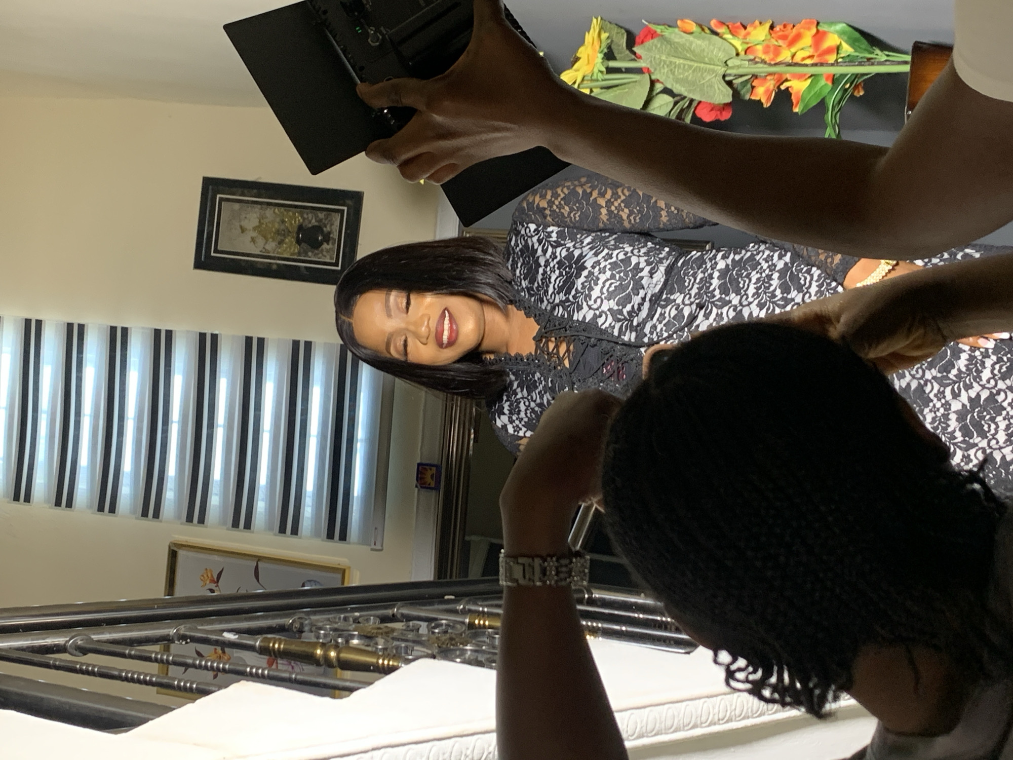 The image captures a scene indoors, seemingly during a photoshoot. A woman with dark hair, wearing a black and white lace dress, is the subject being photographed. Her expression is one of slight amusement, and she is positioned in the center of the frame. The environment includes striped blinds, a framed piece of art on the wall, and another framed picture lower down, visible in the background. The lighting appears to be adjusted by a person holding a reflector or light panel above the subject, and another person is nearby. The presence of equipment and multiple individuals suggest that the location is being used as a studio space. The presence of Nigerian subjects suggest it is indeed in Nigeria, but the location in Jos is not visible from the image.