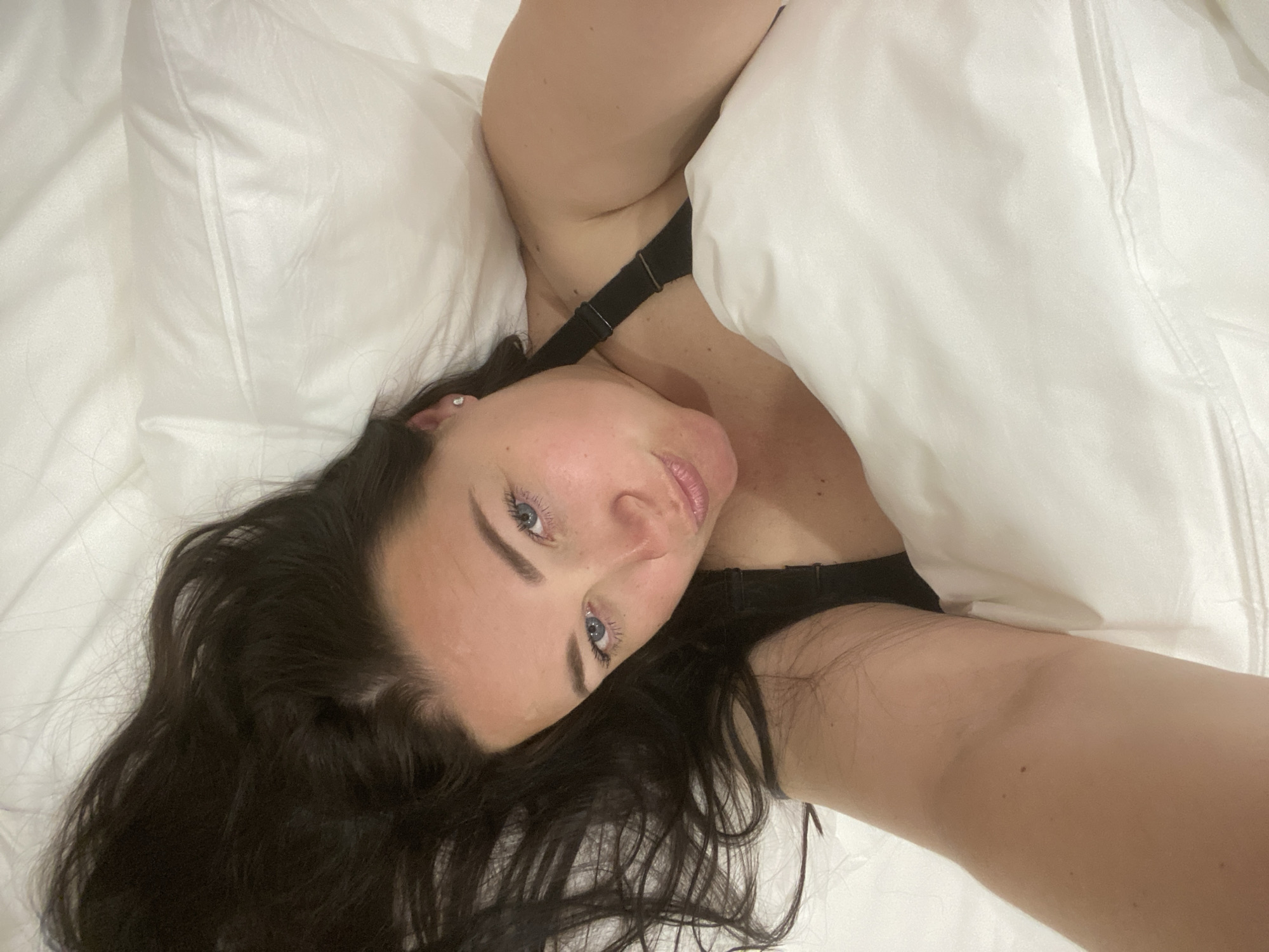 A selfie shows a woman lying in a white bed, partially covered by a white duvet. She has dark hair and blue eyes. A black bra strap is visible. No specific location in Sochi, Russia, can be determined from the image.