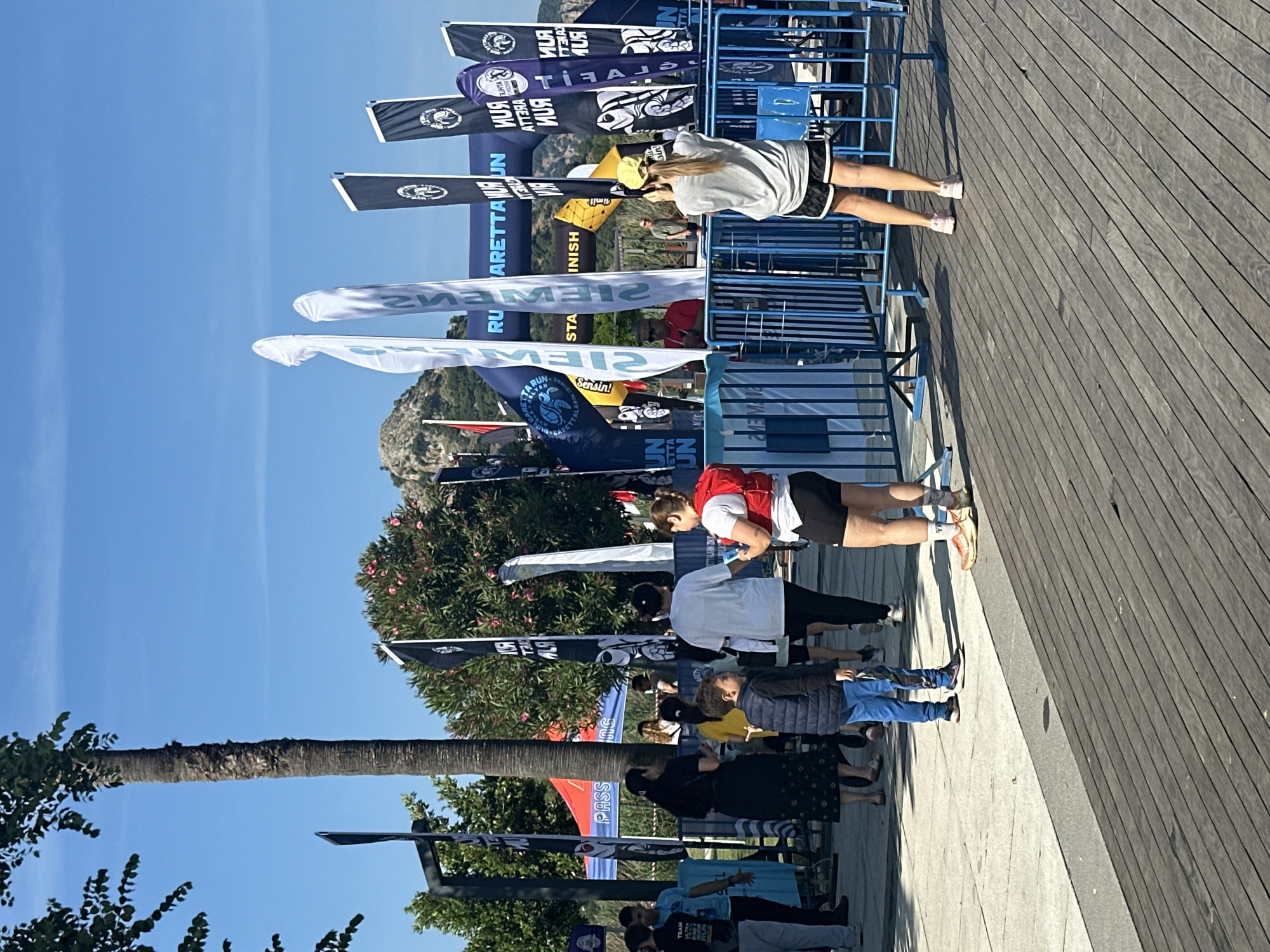 Here is a description of the image:
The image shows a group of people standing alongside a blue metal barricade with various advertising flags. The flags have logos and the text "CARETTA RUN" and "TEAM". Two people are standing near the barricade, a woman in a gray shirt and black shorts, and another person in a white and red shirt and black shorts. A palm tree is visible on the left side. In the background, there is a hill and a clear blue sky. Based on the text on the flags, "Caretta Run," this scene likely occurs at a sporting event, possibly a marathon or race. Based on the text on the flags, "CARETA" this scene likely occurs in Ortaca, Turkey, the location of the Caretta Caretta sea turtle. The people seem to be near a finish line or a designated area for participants and spectators.