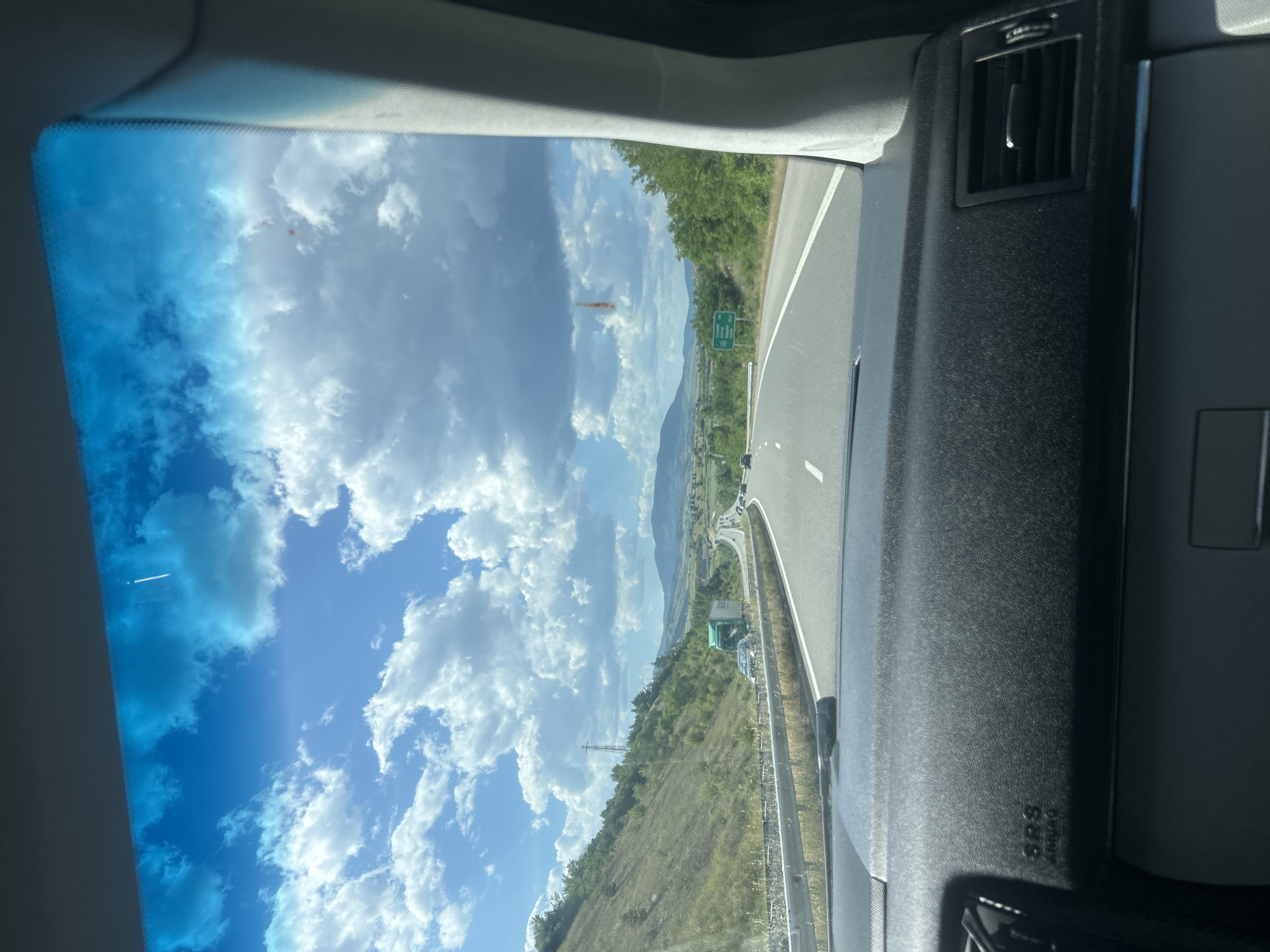 The image, taken from inside a vehicle, shows a highway landscape with a blue sky dotted with white clouds visible through the windshield. The highway has multiple lanes, and other vehicles are visible in the distance. The surrounding area includes mountains and green vegetation, suggesting a rural or mountainous setting. A cell tower is visible. A highway sign is seen in the middle ground. The car's dashboard and air vent are visible in the foreground. The location is likely near Barakovo, Bulgaria based on the presence of mountains and highways in the vicinity.