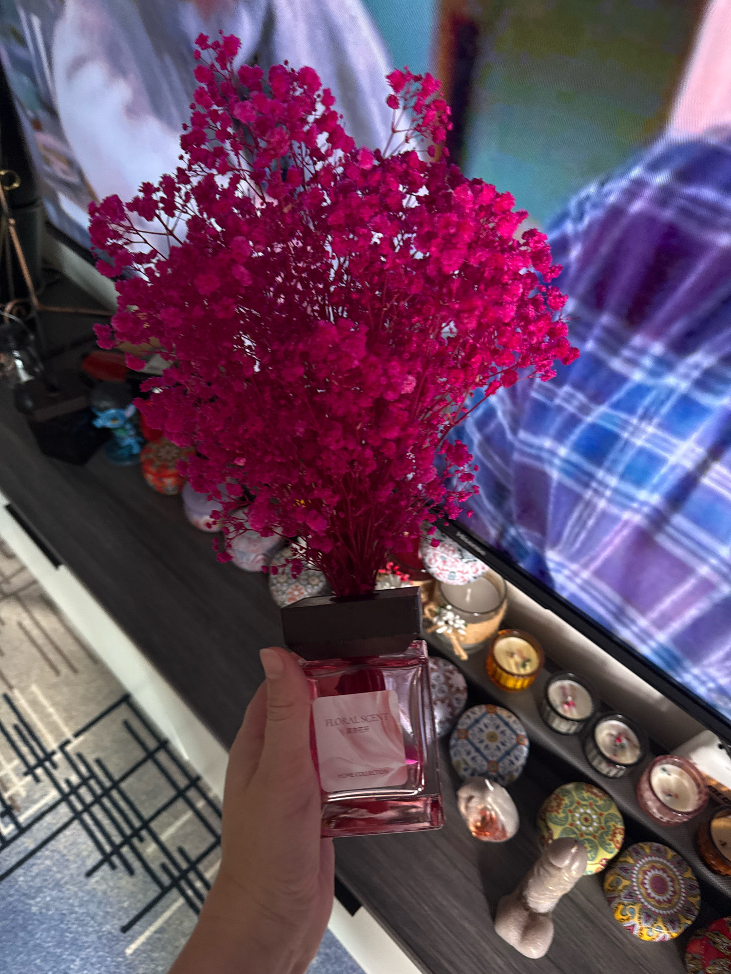 A hand holds a glass perfume bottle filled with pink liquid. The bottle has a label that reads "Floral Scent" and "Home Collection." Behind the bottle, a large bouquet of bright pink dried flowers is displayed. The objects are on a dark gray shelf in front of a television screen showing a person wearing a plaid shirt. To the right of the perfume bottle are several small decorative candles in various colored holders, as well as some small trinkets, including a phallic-shaped object. The scene is set in a domestic environment, likely in Yiwu, China, a city known for its wholesale markets of small commodities.