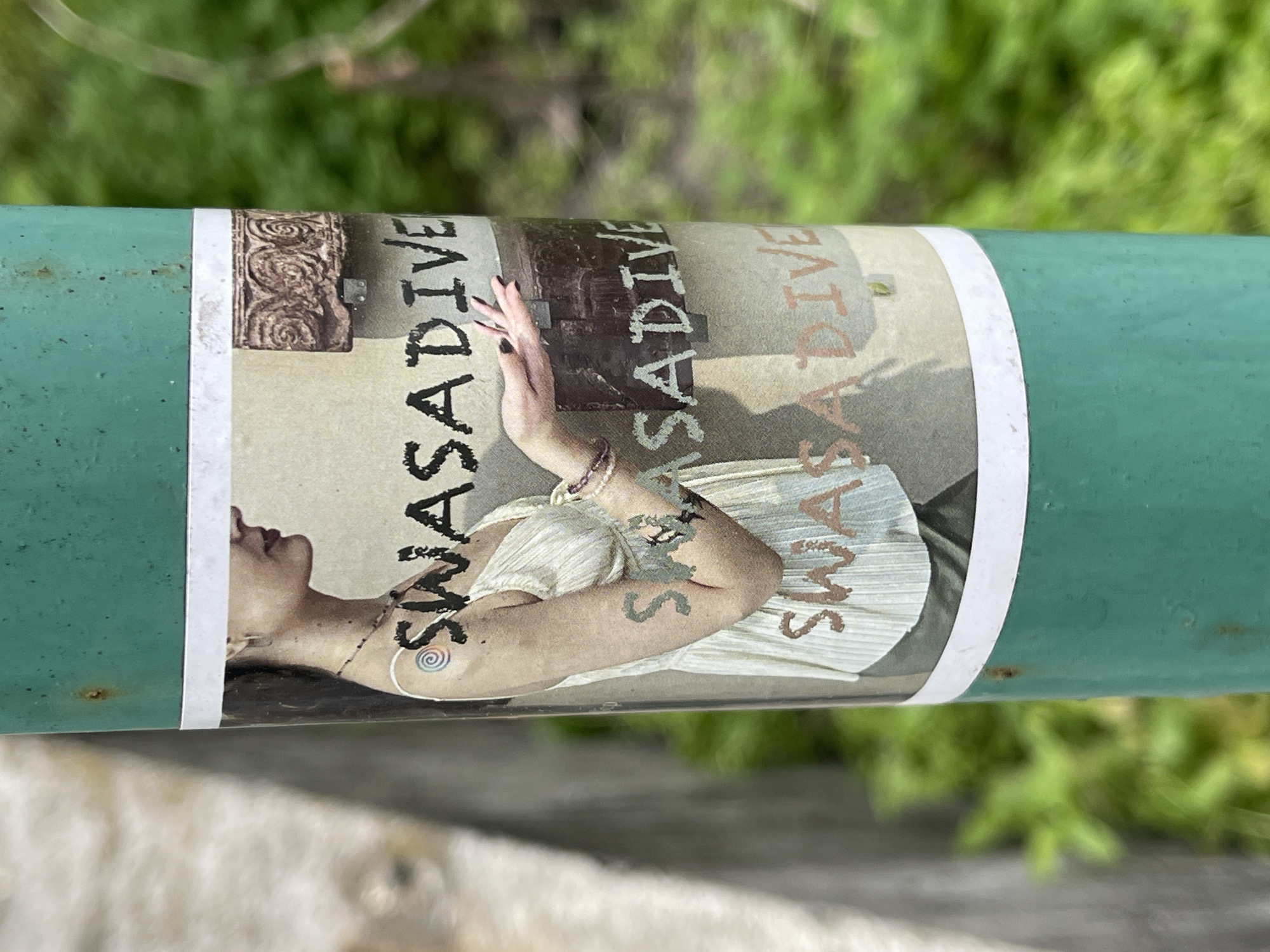 A horizontally oriented image displays a sticker wrapped around a weathered, green metal pole. The pole, speckled with small rust spots, occupies the middle section of the frame. The background, both above and below the pole, is a soft blur of green foliage, indicating an outdoor setting.
The sticker itself features a design with a person and text. The central figure is a fair-skinned individual, likely female, shown from the chest up, with her head tilted back and eyes gently closed or looking upwards. She has dark hair and wears a light-colored, possibly pleated or ruffled, top. Her hands are raised and brought together in front of her chest in a gesture that suggests meditation, prayer, or artistic expression. A bracelet is visible on her left wrist, and intricate body art or tattoos, including a spiral design near her collarbone/shoulder and abstract patterns on her upper arm, adorn her skin.
The background of the sticker reveals several elements: a carved, ornate design with swirling patterns, possibly stone or wood, is visible towards the left. To the right of the figure, a darker, textured area, perhaps resembling an old chest or box, can be seen.
The word "SWASADIVE" is prominently displayed on the sticker multiple times. It appears in varying styles and colors: once in a bold, distressed black font near the figure's left arm, and again in a lighter, faded orange-brown, and a darker grey, often appearing as a layered or ghosted effect. The text has a somewhat rustic or hand-drawn aesthetic.
The image captures the sticker as street art or an advertisement, presented in a public, natural environment.