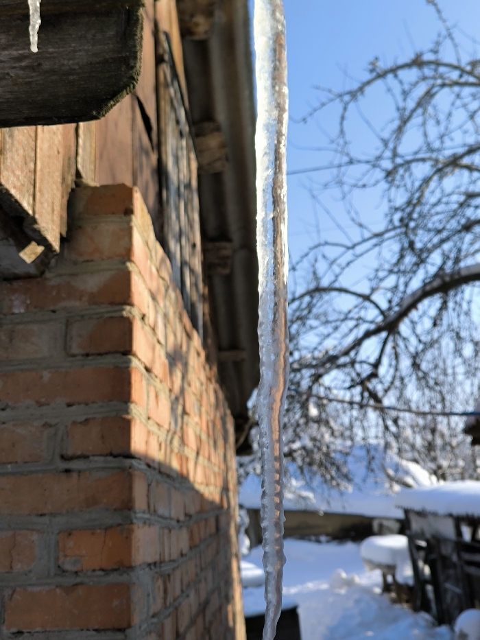 A long, clear icicle, approximately 60-70 cm in length, hangs vertically from the eaves of a brick building in Parkhomivka, Ukraine. The icicle exhibits ridged sections and reflects sunlight. To the left, a smaller icicle dangles from a wooden plank forming part of the eaves. The building's exterior features a wall constructed of red-orange bricks with grey mortar, partially illuminated by direct sunlight on its right side, casting distinct shadows. Above the brick wall, a darker, possibly wooden or metal section, runs horizontally, suggesting a window or upper part of the structure. The background reveals a clear, bright blue sky. Below the sky, bare, dark tree branches, lightly covered in snow or ice, are visible against the distant landscape. The ground in the background is uniformly covered in undisturbed white snow, with what appears to be low, snow-laden structures or furniture situated to the far right. The overall scene depicts a sunny winter day.