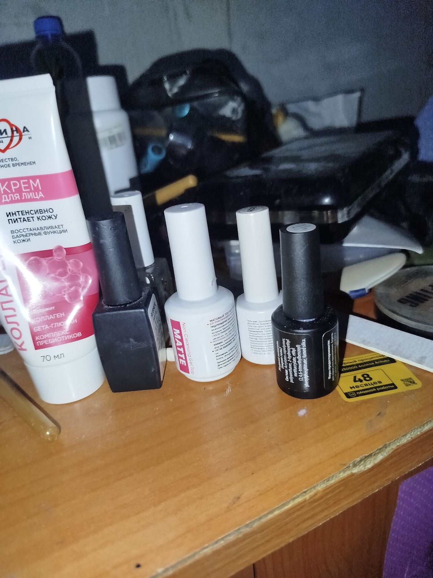 A group of cosmetic products, including a tube of face cream and several bottles of nail polish, are arranged on a wooden surface. The face cream tube is white with pink and red lettering, and it has "КОЛЛАГЕН" and "70 мл" visible. To the right of the cream, there are four bottles. The first bottle from the left is black with a black cap, and appears to be a nail polish. Next to it is a white bottle with a white cap, also a nail polish, with "MATTE" and "Non Cleansing Gel Top" visible on its label. To the right of this is another white bottle with a white cap, similar in size and shape to the "MATTE" bottle. The last visible bottle is black with a black cap, and has text in Russian and English, including chemical names. To the far right, partially visible, is a yellow sticker with the number "48" and Russian text. The background is dark and out of focus, showing what appears to be other objects and possibly a dark plastic bag. The lighting is dim, suggesting an indoor environment. The scene is consistent with a personal vanity or a retail display. There is no indication of the city of Zavodoukovsk, Russia, in the image.