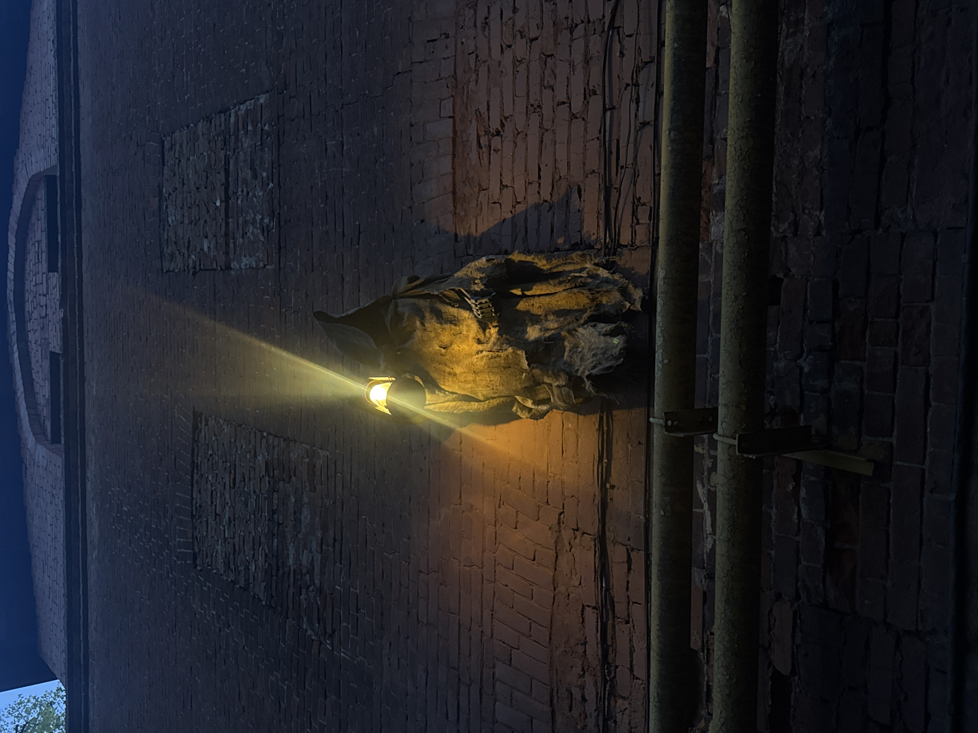 The image shows a brick wall with pipes running vertically along its right side. Attached to the wall is a light fixture casting a yellowish glow. Above the light, the wall appears to have some covering or burlap-like material. Parts of the brickwork look like filled-in windows. The location is identified as Nizhny Novgorod, Russia.