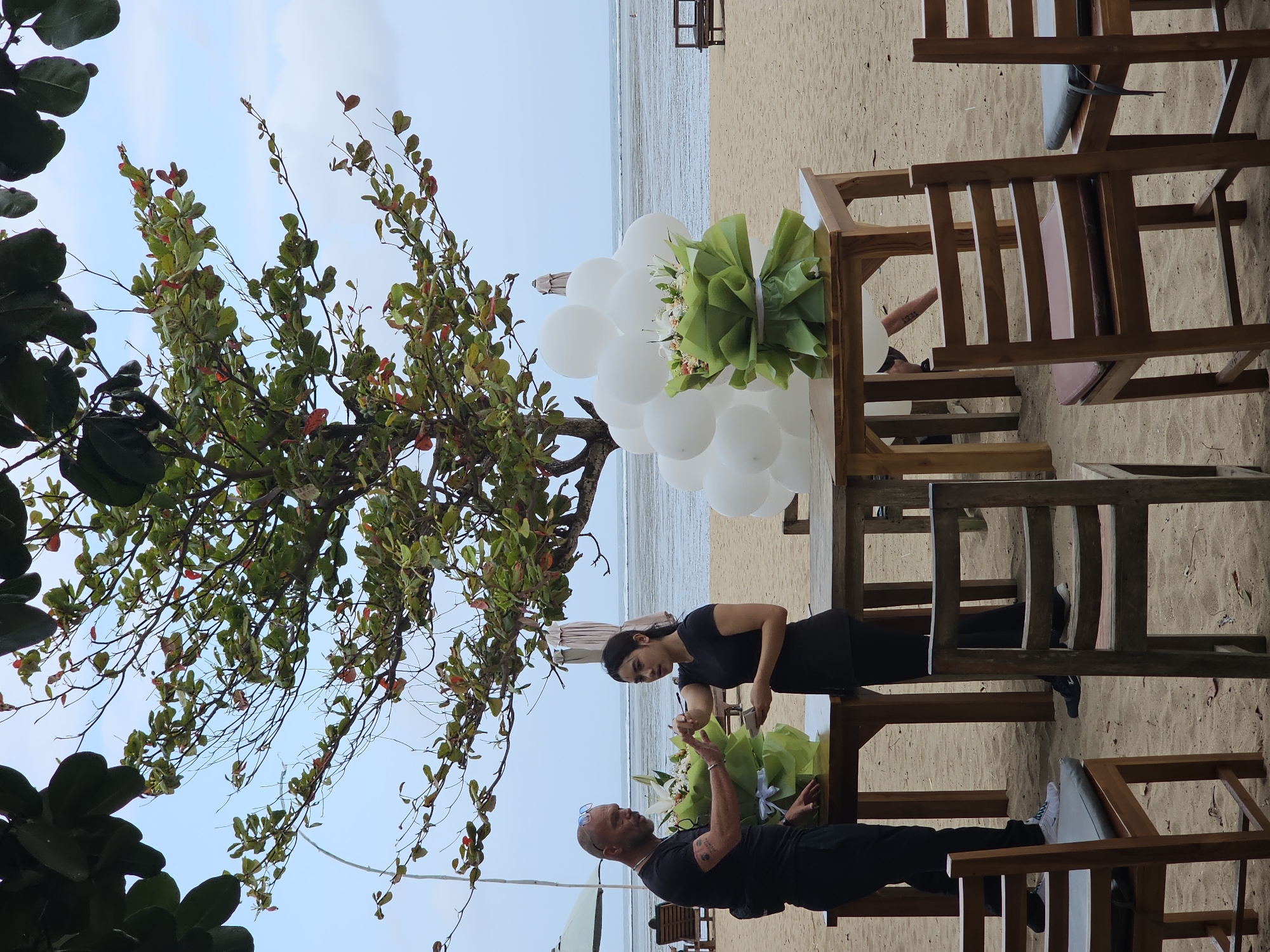 The image depicts a beach scene in Denpasar, Indonesia, with a man and a woman standing behind a table laden with floral arrangements and white balloons. The man, wearing a black t-shirt and black pants, has sunglasses on his head and is looking at the woman. The woman, in a black t-shirt and black pants, is holding a small object, possibly a device or a box, and is interacting with the man. In front of the table, several wooden chairs are placed on the sand. Behind the table, the ocean stretches to the horizon under a cloudy sky, with a tree providing shade over the setup. The overall atmosphere suggests preparation for an event, possibly a wedding ceremony given the romantic setting and floral decorations.