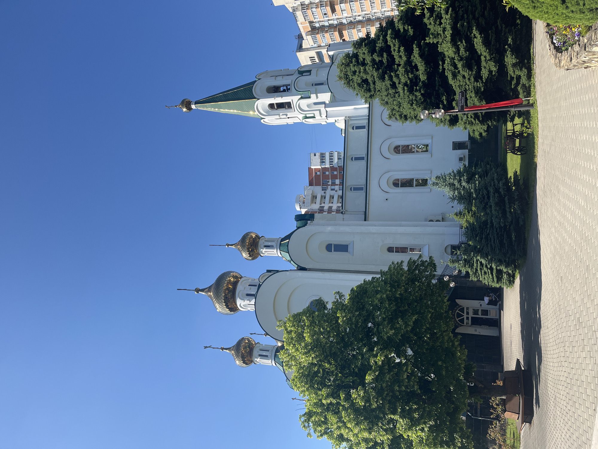 The image presents the Cathedral of the Holy Great Martyr Catherine (Ekaterininsky Cathedral) in Krasnodar, Russia, under a clear, bright blue sky. The cathedral features a white facade, multiple green roofs, and five visible golden onion domes, each surmounted by an Orthodox cross. A tall, slender bell tower with a green, pointed spire and a golden cross is positioned on the right side of the main structure, incorporating arched openings for bells.
The foreground consists of a light grey paver walkway or plaza. On the left, a large deciduous tree with full green foliage and a circular wooden bench at its base partially obscures the lower section of the church's entrance. Several dark green coniferous trees are distributed across the church grounds, particularly on the right side. A decorative wooden cart and a lamppost with a directional sign (displaying "ЦЕРКОВНАЯ ЛАВКА" - Church Shop, and "ТРАПЕЗНАЯ" - Refectory in Cyrillic) and a red banner are visible on the right.
One individual in light-colored clothing is partially visible near the main entrance on the left, pushing what appears to be a stroller or small cart. In the background, modern multi-story residential buildings with facades in white, orange, and red tones are visible, indicating an urban environment. The scene is brightly illuminated by direct sunlight, casting distinct shadows.