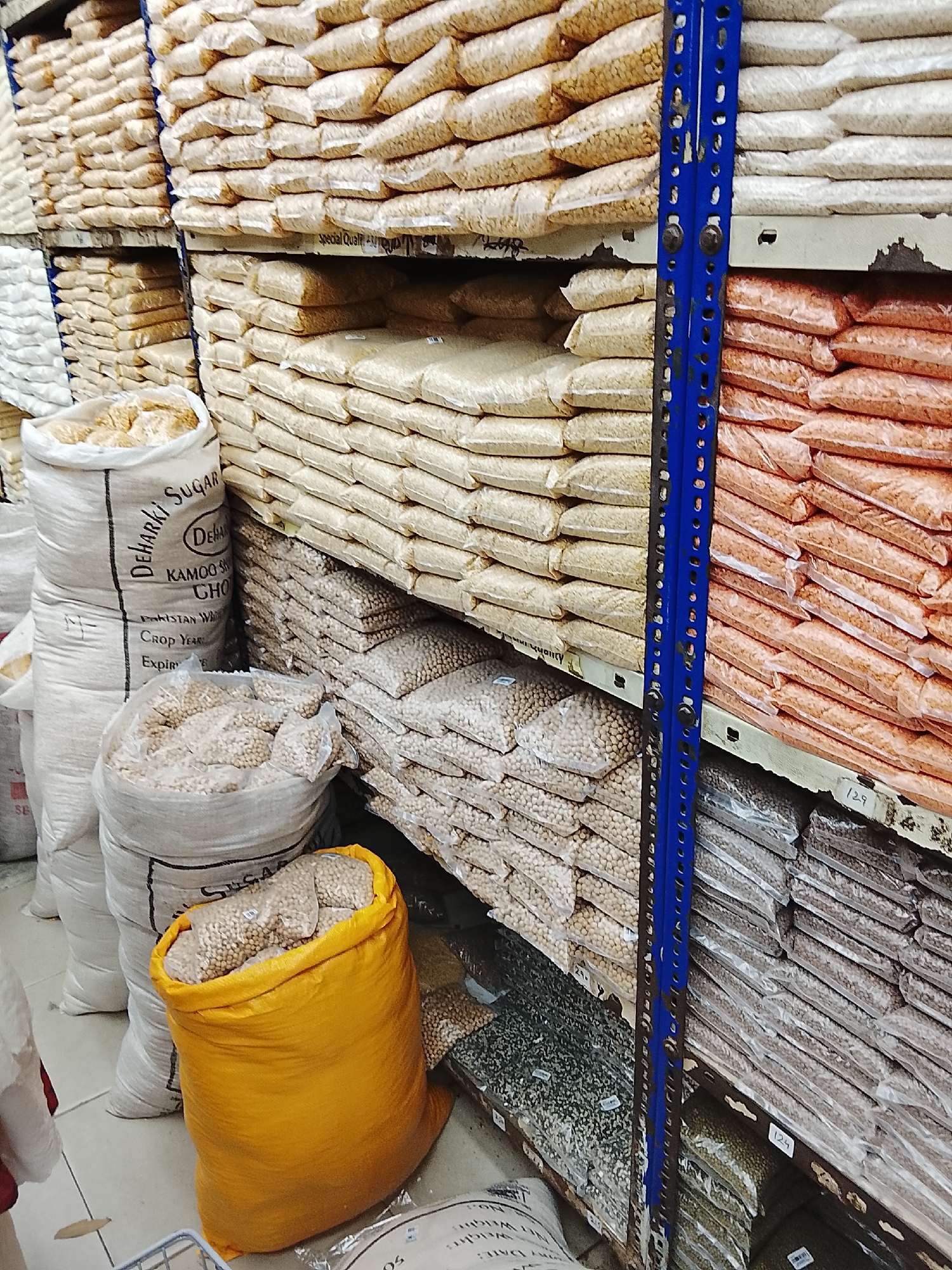 The image captures the interior of a storage or retail space in Gulgasht Colony, Pakistan. The scene features shelves stacked high with clear plastic bags and large sacks of what appear to be beans, lentils, and grains. The products are arranged in rows, demonstrating an effort at organization.
A distinctive bright yellow sack stands out in the foreground, positioned in front of several white sacks labeled with the word "SUGAR." A blue, perforated metal shelf support is visible along the right side, emphasizing the industrial or commercial setting.
The lighting is somewhat dim and the overall impression is one of a packed, utilitarian storage area, typical of a small retail or storage space.