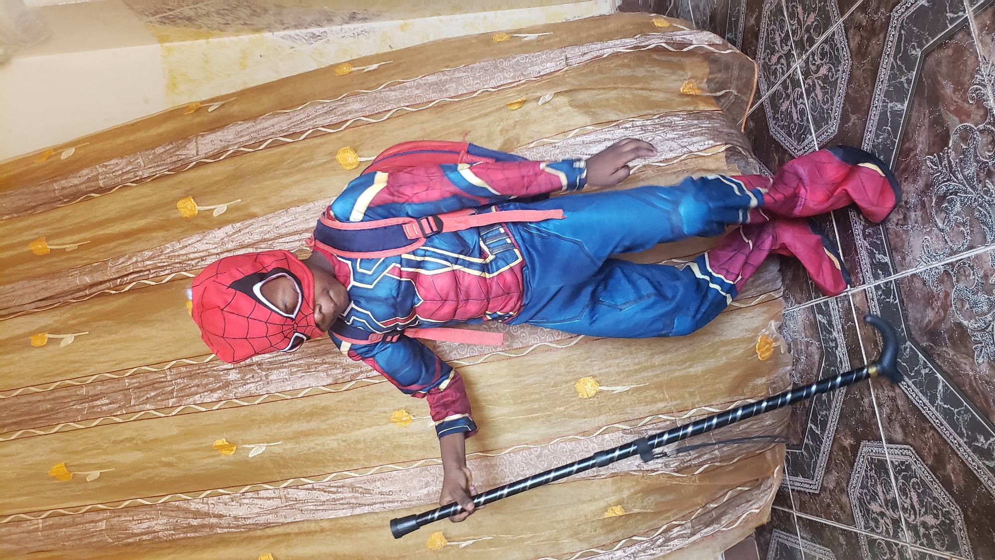 A child in a Spider-Man costume lies on a bed covered with a patterned, gold-colored bedspread. The child is wearing a red and blue Spider-Man mask and suit with attached red boot covers. The child lies diagonally across the bed, grasping a black cane with a curved handle in the lower left corner of the image. Part of a tiled floor is visible along the right edge of the bed. The location is Narayi, Nigeria.