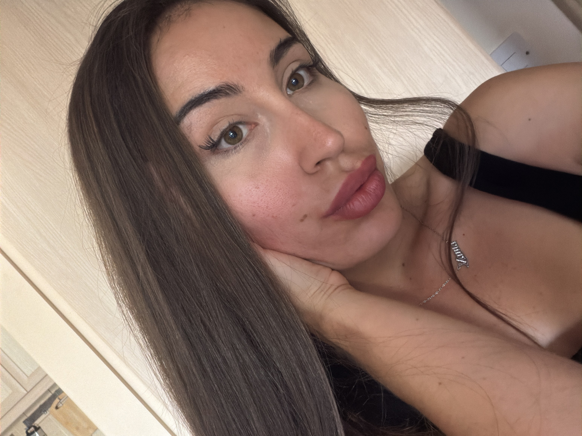 A close-up, medium shot captures a young woman with long, dark brown hair taking a selfie. Her left hand is resting on her cheek, and she is wearing a black top and a silver necklace with the name "Lana" in cursive. The background appears to be an indoor setting with light-colored wooden paneling. The lighting is soft and natural, suggesting daylight. There is no indication of the specific location in Tersefanou, Cyprus.