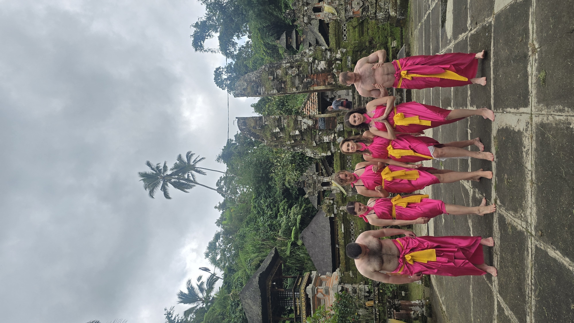 The image depicts a group of six people in the city of Tegallalang, Indonesia, in an outdoor setting that includes traditional Balinese structures and tropical vegetation. Two shirtless men and four women are posing in a line. They are all wearing fuchsia sarongs with yellow sashes tied around their waists. The ground is paved with square stones, and behind the group are moss-covered Balinese temple structures and dense tropical trees. The sky is overcast.