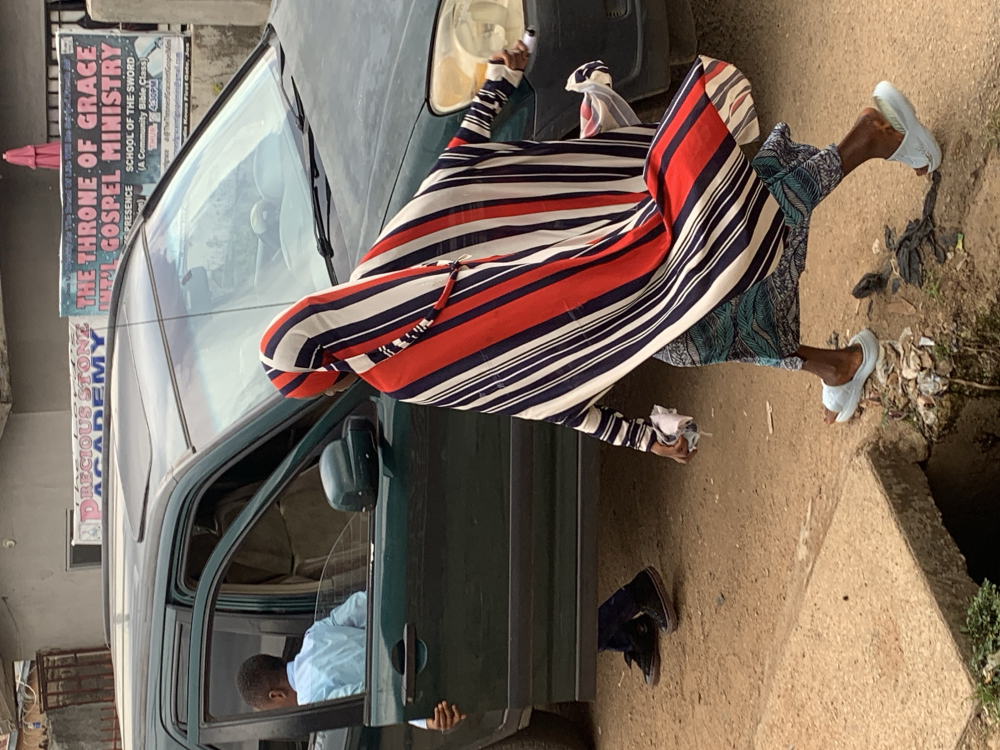 The image depicts a street scene in Jos, Nigeria, with a dark green SUV in the foreground. A young man wearing a light blue shirt is seated in the driver's seat with the driver's side door open. He appears to be looking away from the camera. A person wearing a colorful striped abaya (red, white, and navy blue) is walking past the SUV from right to left. The abaya is flowing, suggesting movement. This person is also wearing a patterned skirt and light blue slides. In their right hand, they are holding a white object, possibly a piece of paper or a tissue. To the left of the SUV, another person, likely male given the black sneakers and dark pants, is standing with his left hand on the open car door, appearing to be leaning into the vehicle. In the background, banners are visible. One banner reads "THE THRONE OF GRACE INT'L GOSPEL MINISTRY," and another partially visible banner reads "PRECIOUS STONE ACADEMY." The ground is dirt and appears to be a street, with some debris visible near a concrete culvert or drainage ditch on the left. The overall scene suggests a moment of interaction or transition related to the vehicle.