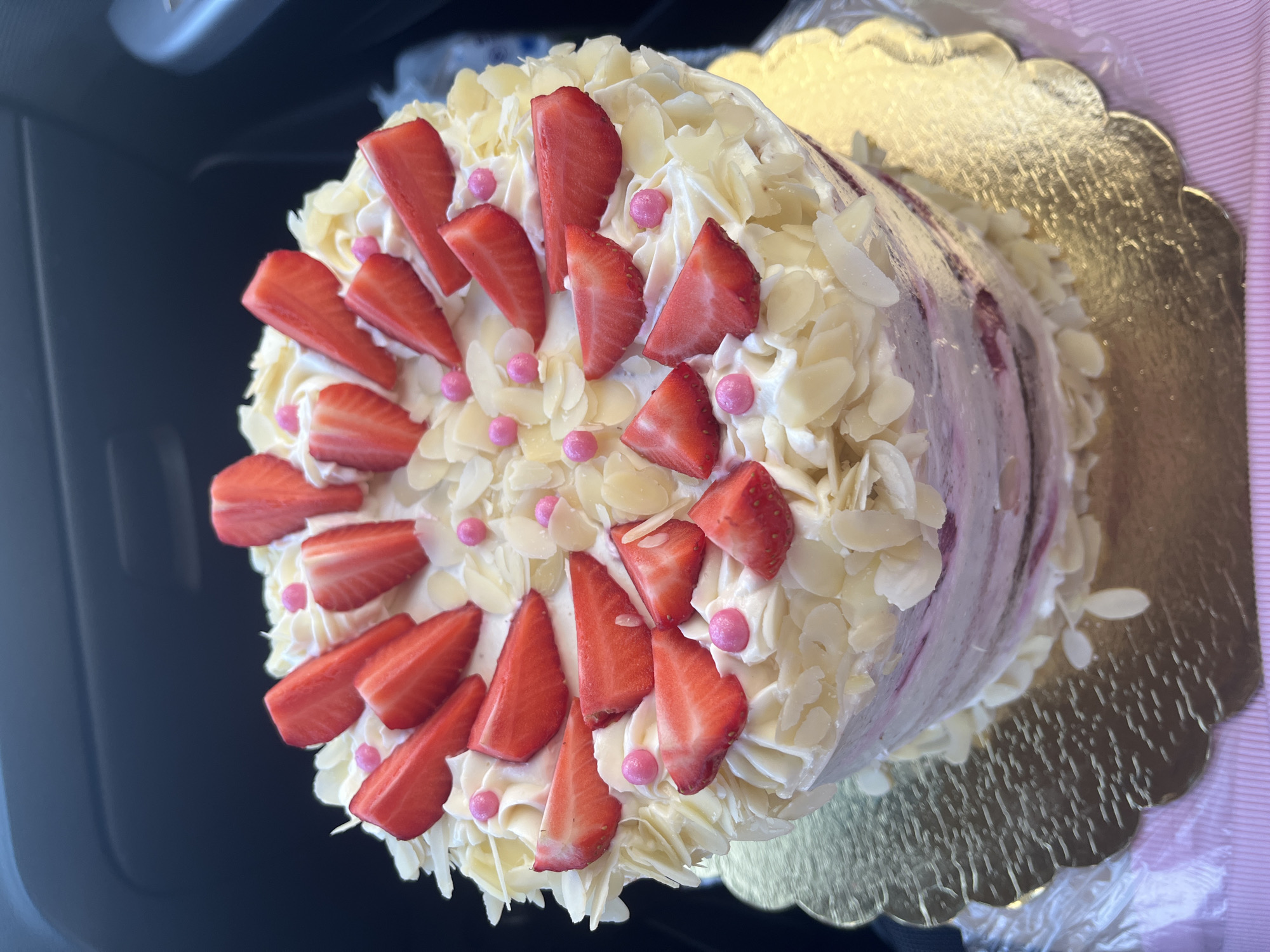 A visually prominent, round, multi-layered cake is centered in the frame. The cake is topped with white whipped cream or frosting, arranged in swirls. Scattered across the top and cascading down the upper sides are numerous red strawberry slices, mostly halved longitudinally. Interspersed among the strawberries and cream are numerous irregular white almond slivers or flakes, and small, spherical, glossy pink sprinkles. The sides of the cake exhibit a partially exposed, light pink or purple marbled texture beneath the white frosting, suggesting a semi-naked cake design. The cake rests on a golden, scalloped-edge cake board. Below the cake board, a textured, light pink surface is visible, partially covered by clear plastic wrapping. The background is dark and out of focus, revealing indistinct shapes consistent with the interior of a vehicle, possibly showing elements of a dashboard or door panel. No individuals are visible. The specific location in Krasnodar, Russia, cannot be determined from the visual information provided in the image.