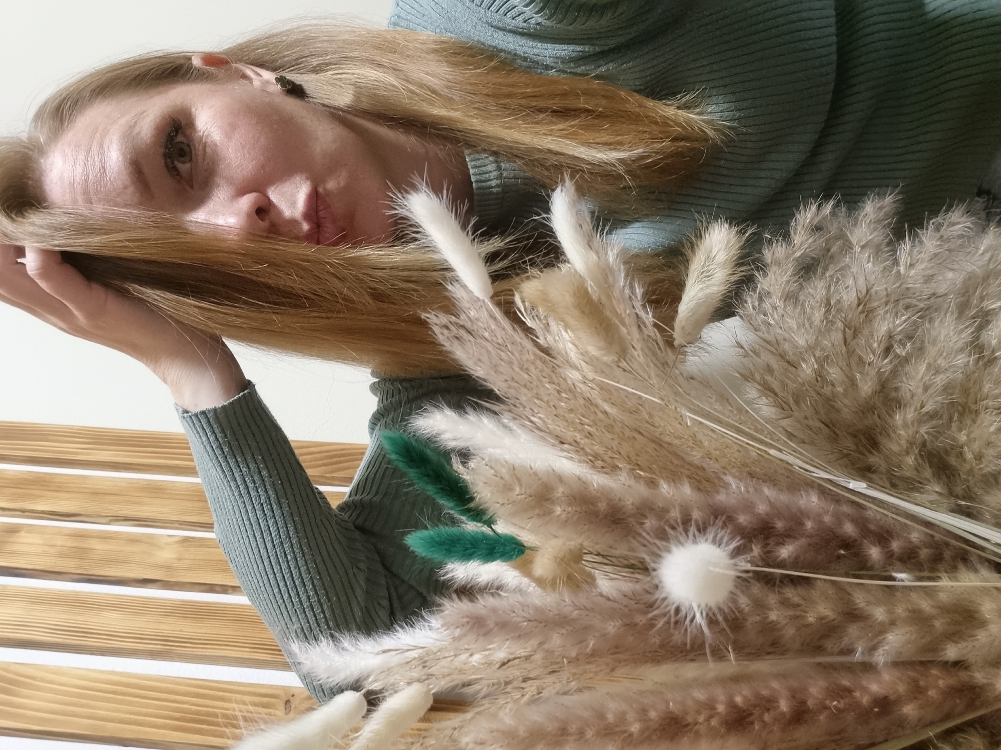 A woman with fair skin and blonde hair is captured in an angled, close-up shot. She is wearing a sage green turtleneck sweater and has her left hand near her hair. A bouquet of dried grasses and flowers, including a few dyed green, is held in front of her, partially obscuring her body. The background includes a wooden plank surface of light brown color. The image appears to be taken indoors. There is no specific evidence in the image that would pinpoint the location as Chaikovsky, Russia.