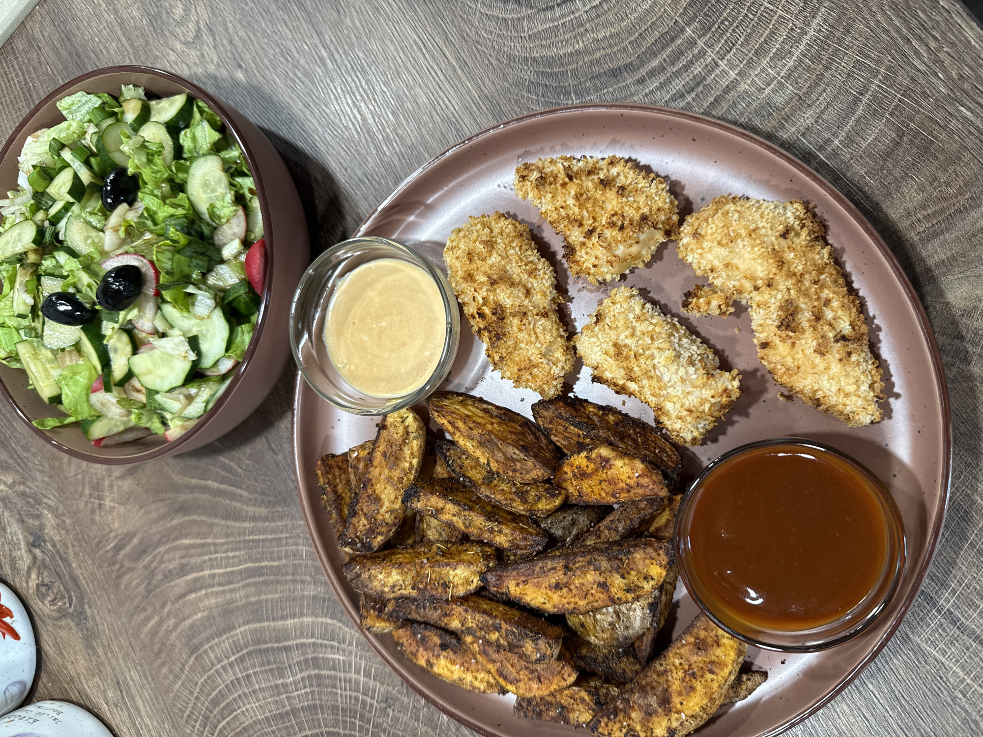 The image displays a prepared meal set on a dark, visibly grained wooden table. In the foreground, an oval, brownish-mauve plate contains the main components. On the left side of the plate, a pile of seasoned potato wedges, characterized by their dark golden-brown, somewhat charred appearance and visible seasoning, is presented. To the right of the potato wedges are several irregularly shaped pieces of breaded chicken, featuring a light golden-brown, visibly crispy coating. Two small, clear glass bowls of dipping sauces are placed on this main plate: one in the bottom left contains a reddish-brown, viscous sauce, and another towards the top right holds a lighter tan, creamy sauce. In the upper right background, a round bowl, matching the color of the main plate, is filled with a fresh green salad comprising chopped lettuce, sliced cucumbers, and sliced red radishes, garnished with three black olives. Partially visible in the upper left background are the rims of two ceramic mugs, one featuring text that reads "KITCHEN" and a floral design, and the other displaying a red chili pepper. No people are visible in the scene. The lighting suggests an indoor setting, likely a home dining area.
