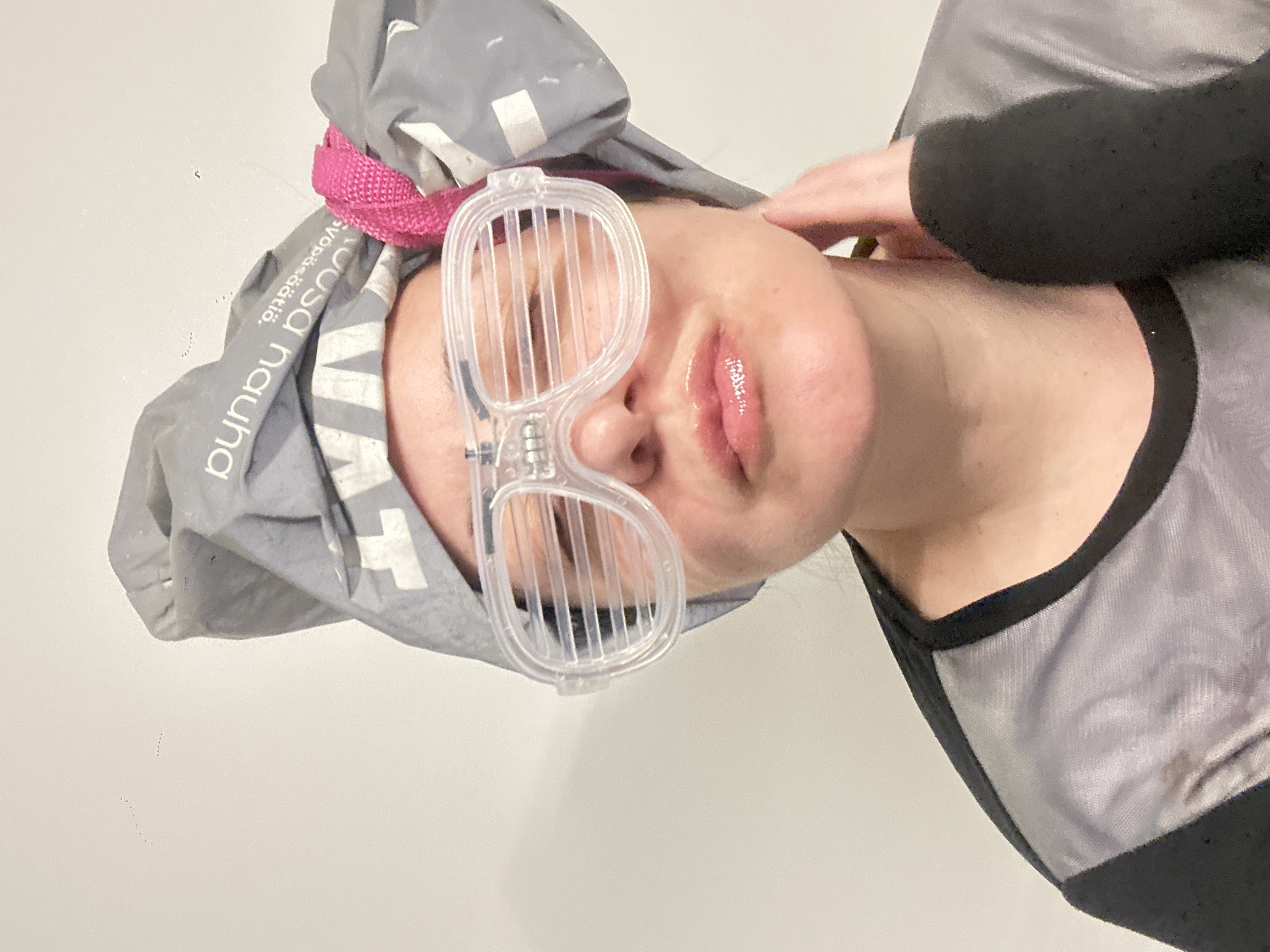 A close-up, rotated portrait features a person wearing clear plastic pinhole glasses and a grey fabric head covering. The individual looks towards the viewer with slightly parted, glossy lips. The pinhole glasses, designed with vertical slits, obscure their eyes. A bright pink strap or band secures the grey head covering, on which partially visible text reads "Suosa nauha" and "svöpsä.dätlõ.". The person wears a grey and black garment, and one hand is gently placed on the side of their neck. The background is a plain, light-colored surface, likely a wall. The overall scene suggests a personal wellness or therapeutic activity, possibly involving vision correction exercises or a relaxation treatment like a sauna, given the nature of the headwear. The image does not provide cues for time of day or outdoor location.