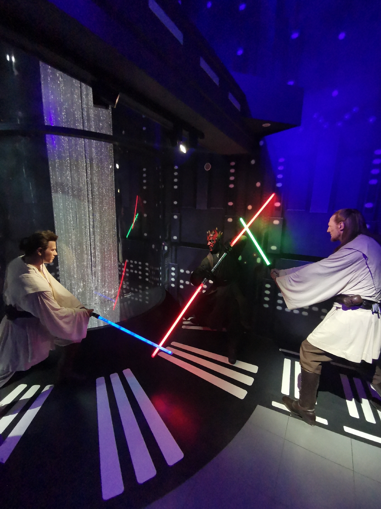 The image shows a Star Wars diorama, featuring wax figures of Obi-Wan Kenobi, Qui-Gon Jinn, and Darth Maul in a lightsaber battle scene. The figures are positioned in front of a backdrop simulating a spaceship interior. The floor has a black and white striped pattern, mimicking a futuristic setting. The figures are holding lightsabers with red, blue, and green blades. The lighting in the diorama uses blue and red hues, creating a dramatic effect. The location is likely a display at Madame Tussauds in London, United Kingdom.